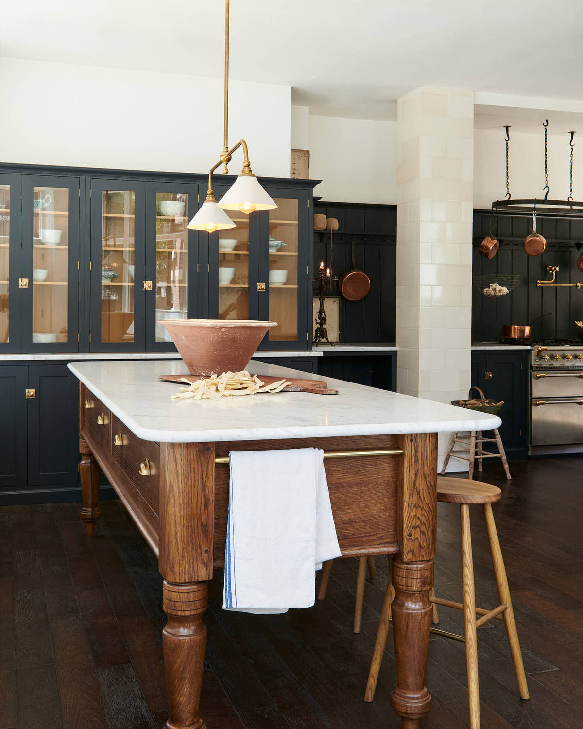 A large pantry blue shaker kitchen features a sink and cooker run with Carrara marble worktop, countertop cupboards with Scullery yellow interior, and a wide Heirloom dairy table. Beautifully decorated with artisan ceramics, copper pans, deVOL Heirloom gaselier lights, a pot hanger, an aged brass pot filler tap, and Helen Stools, the kitchen presents a thoughtfully curated mix of collected pieces that fit together effortlessly.