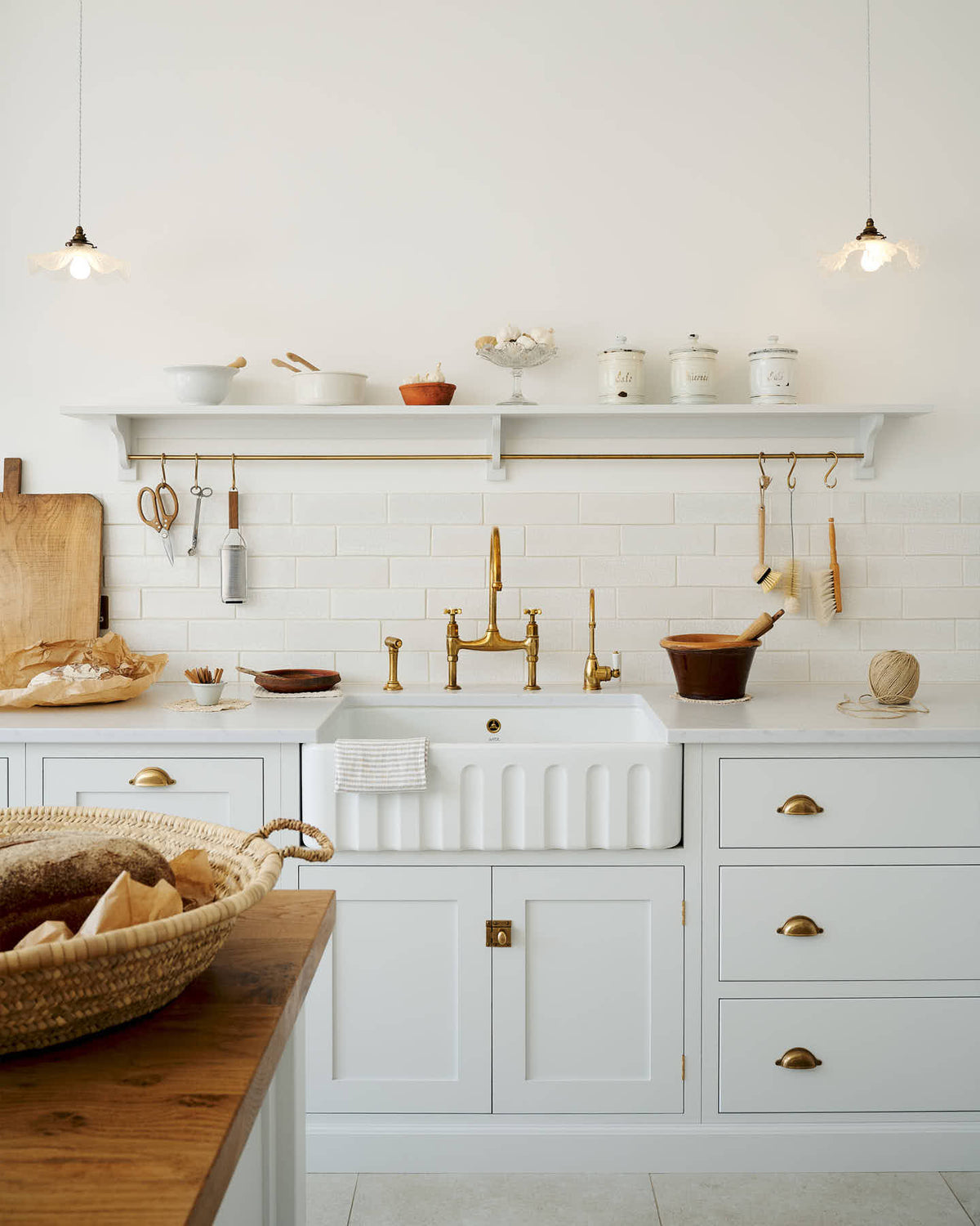 A closer look at a Damask off-white shaker kitchen featuring a solid oak island and Carrara marble worktops on the sink and cooker runs. The sink run includes a single fluted Belfast sink with an aged brass Ionian tap. Crackle metro splash back tiles adorn the walls, while a Shaker shelf with aged brass rails holds essentials like scissors, scrubs, and a grater.