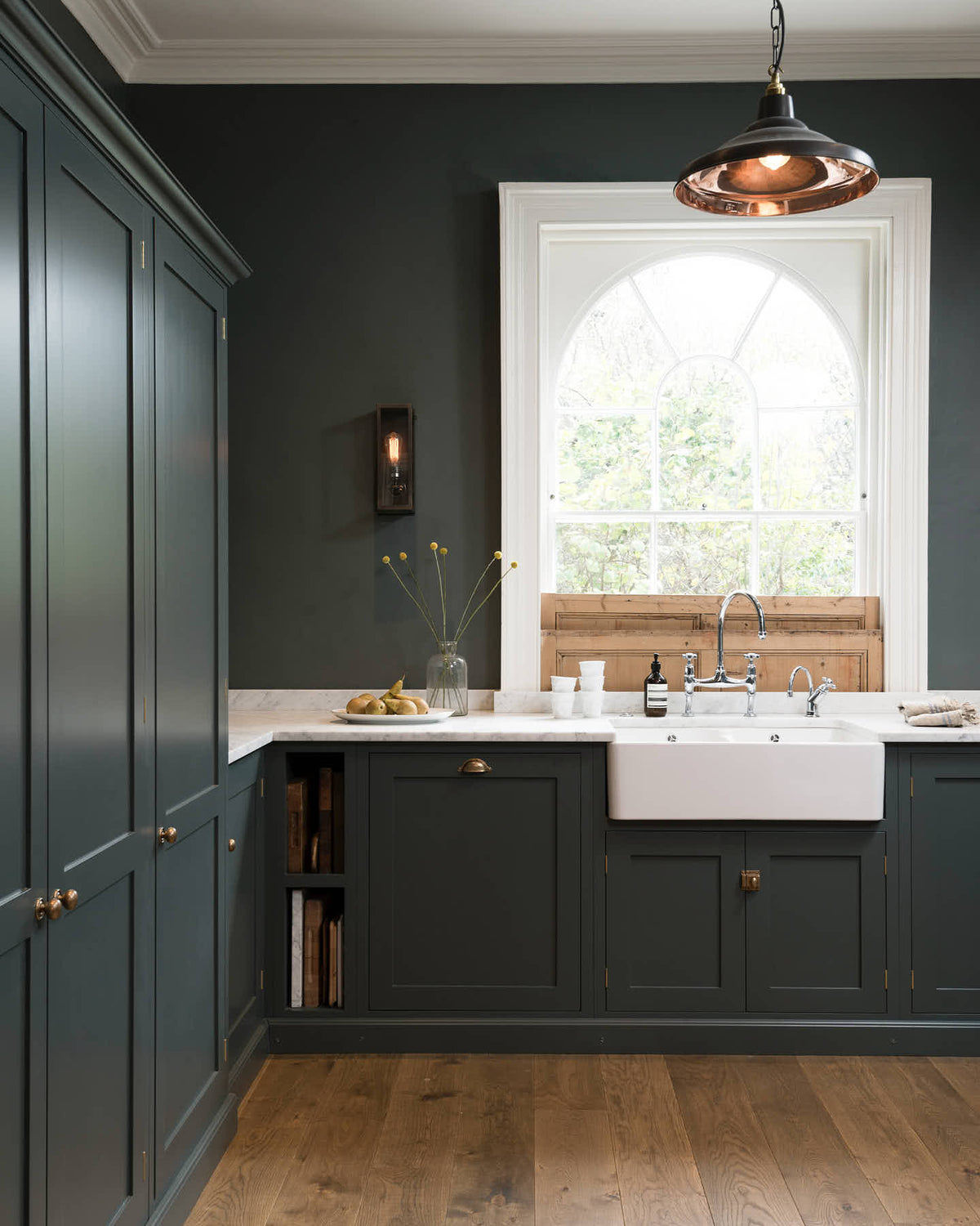A charcoal-black, classic shaker kitchen features a U-shaped sink and cooker run with Carrara marble worktop, a chrome Ionian tap, open countertop cupboards, a prep table with a solid oak worktop and a bank of fitted pantry cabinets. Beautifully decorated with a large vintage ceramic bowl, steampunk wall lights, a copper pendant light, fresh pears, and the original Georgian wooden sliders, the family kitchen strikes a wonderful balance between modern style and timeless character.