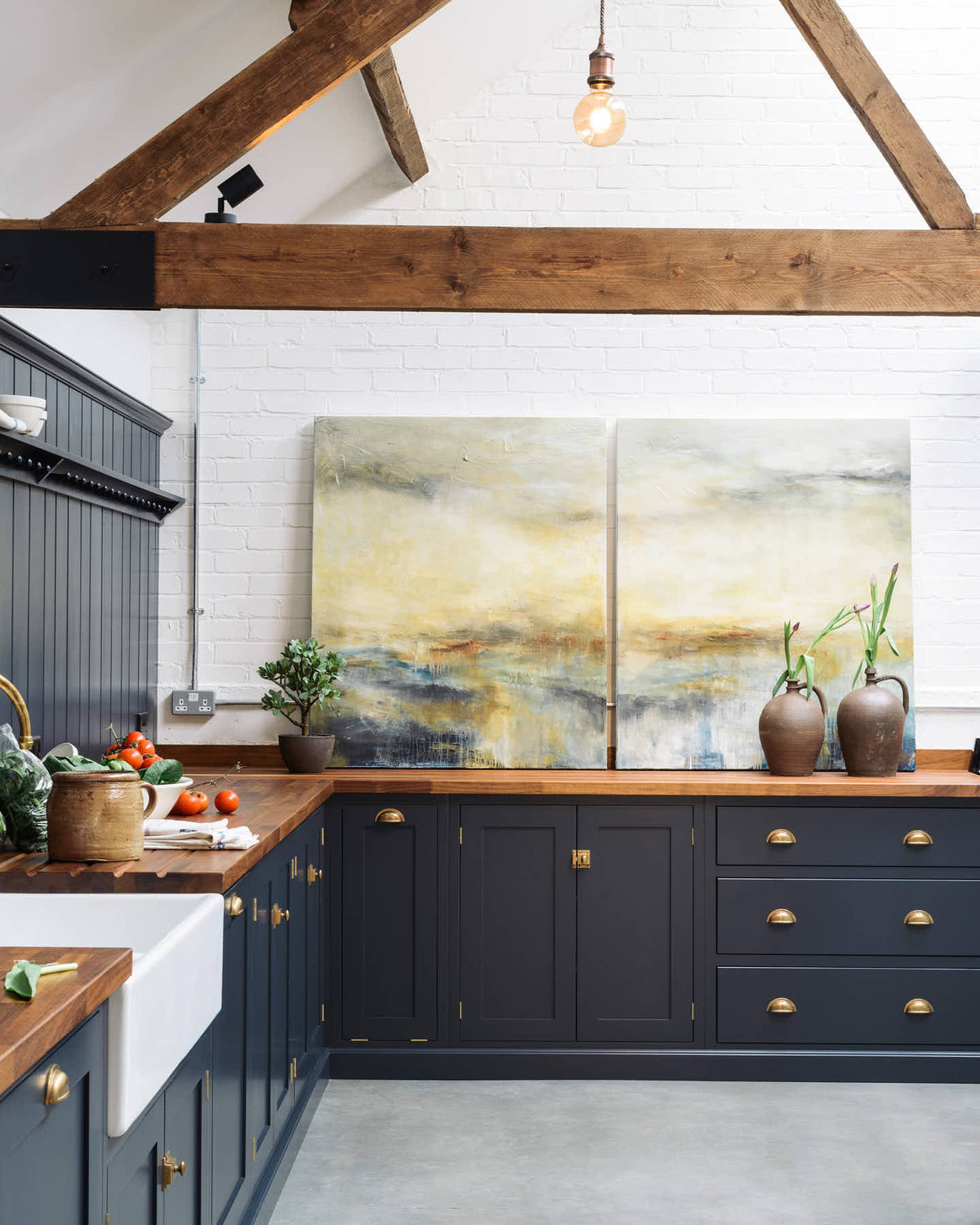Nestled between an L-shaped cooker and sink run and a large white linen kitchen island topped with a stave hardwood worktop, the spacious pantry-blue Shaker kitchen has transformed a large, renovated cattle shed. Beautifully decorated with aged brass taps, Helen stools, lucious green vegetables, vintaged artisan ceramics, lilac tulips and abstract paintings.