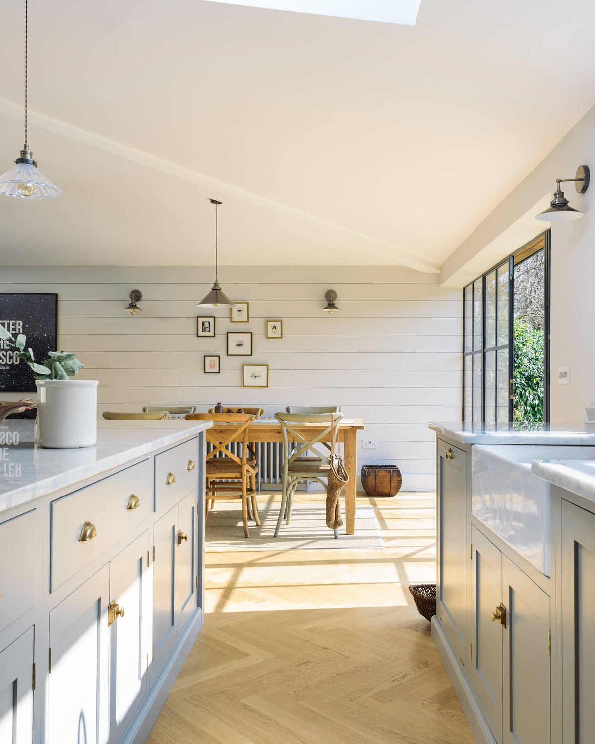 Nestled between the L-shaped sink and cooker run with a Carrara worktop and a kitchen island in a spacious and contemporary stone grey Shaker kitchen, is a single Belfast sink with an aged brass Ionian tap. Adjacent to this setup is a grand dining room featuring a long, solid oak dining table and chairs.