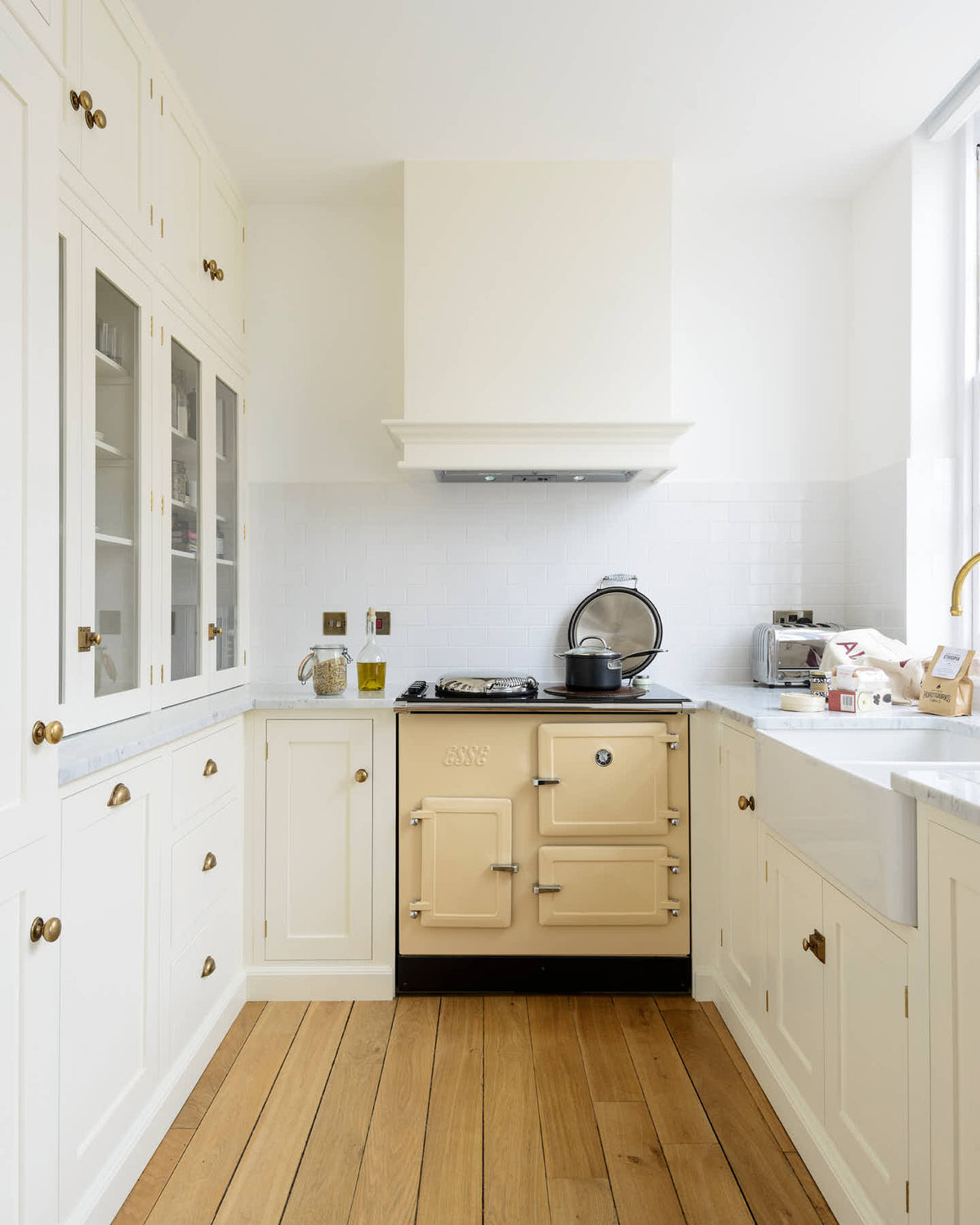 This small but functional kitchen features a white shaker design, complete with an L-shaped layout that includes a cooker and sink run topped with Carrara marble. It boasts a shaker-style canopy, glazed cupboards, drawers, and a white porcelain splash back. Additionally, there is a pantry and extra storage cupboards. The pale wooden plank flooring, aged brass handles, and cream gas stove add warmth to this bright and simple kitchen.
