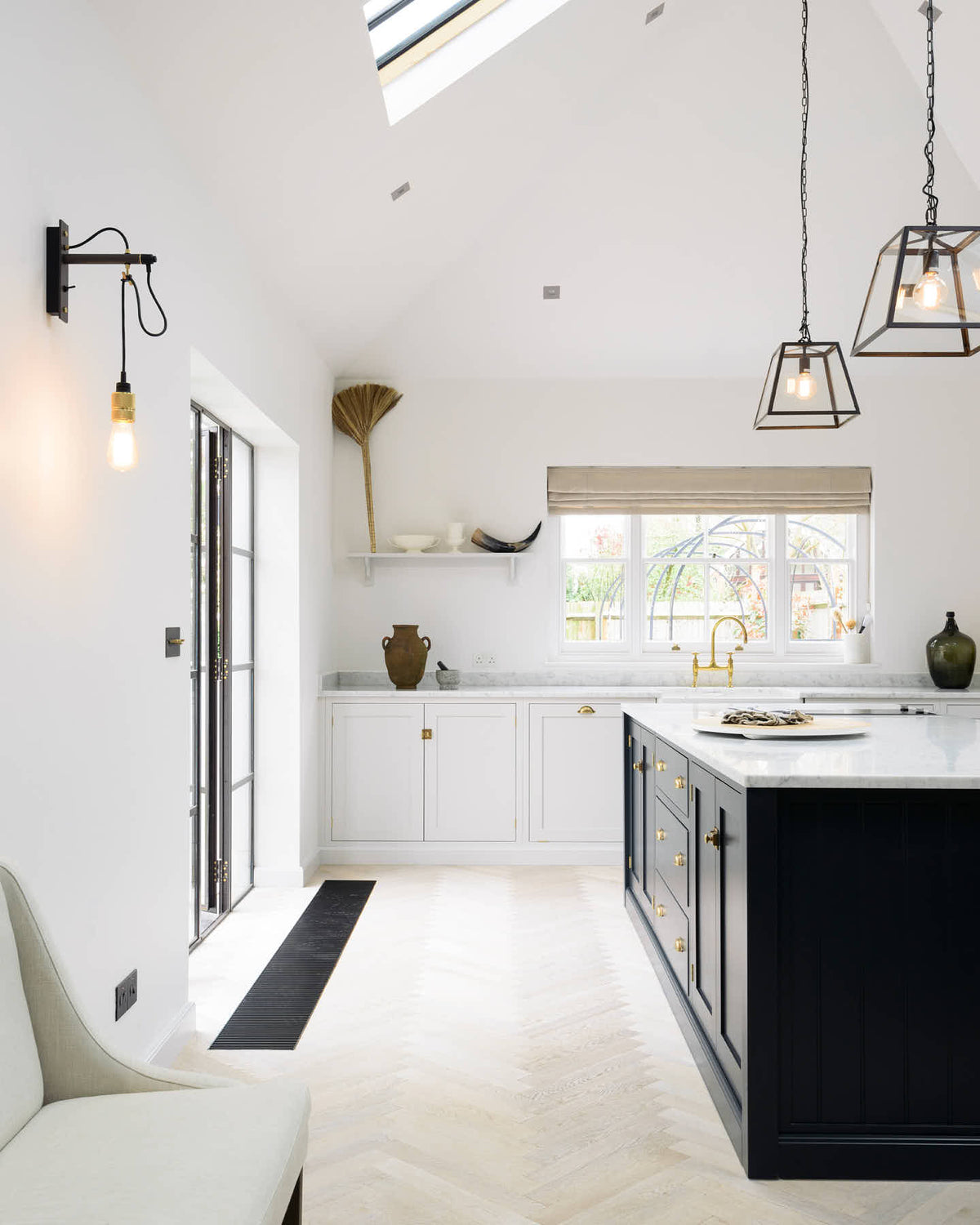 Nestled between an off-white L-shaped sink run with a double Belfast sink and an aged brass Ionian tap, and a pantry-blue kitchen island with a Carrara marble worktop, is a spacious and artisan Shaker kitchen. Cleverly decorated with steel-framed hanging lights, large glasses with narrow necks, vintage ceramics, and eclectic items serving as artistic displays, it creates a simple yet intriguing kitchen space.