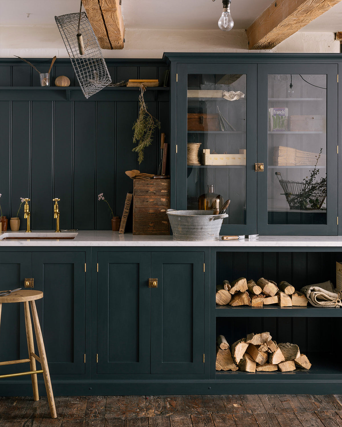 A closer look at the sample pantry blue shaker kitchen’s sink run features a shelf, countertop glazed cupboards, and a mix of closed and open base cabinets. The Carrara marble worktop holds a copper single sink with aged brass Mayan taps. Wooden and metal accents—including firewood, a metal bin, and a wooden toolbox—add rustic charm.