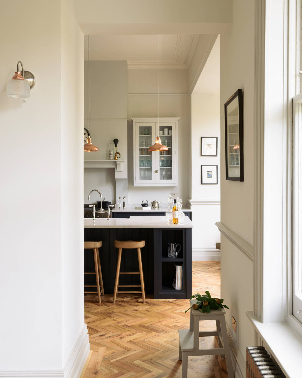 The pantry blue shaker kitchen features creamy silestone worktops. Off-white walls frame two pale wall cupboards flanking the chimney breast. The central island features a sink on one side and a breakfast bar with wooden stools on the other. The kitchen and dining room have been combined into one large space, with a central column providing subtle separation.