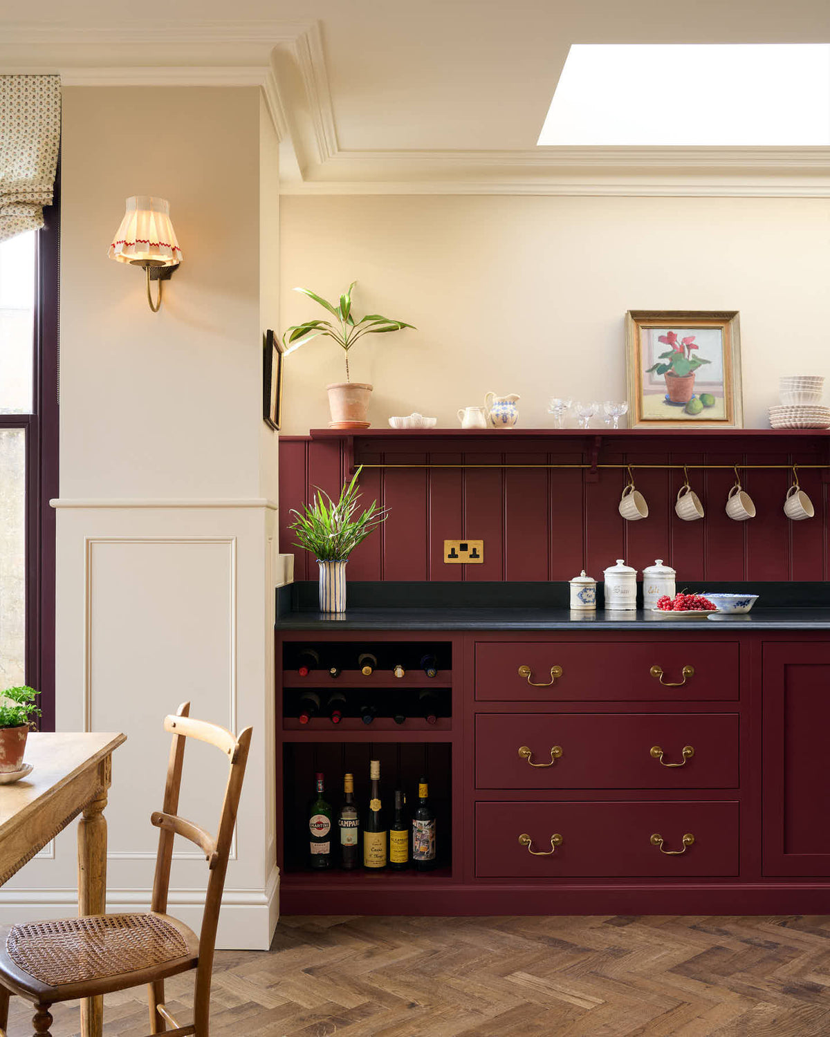 A straight-on view of a refectory red Shaker kitchen with parquet wood flooring, cream walls, a black granite worktop and red tongue and groove panelling above.