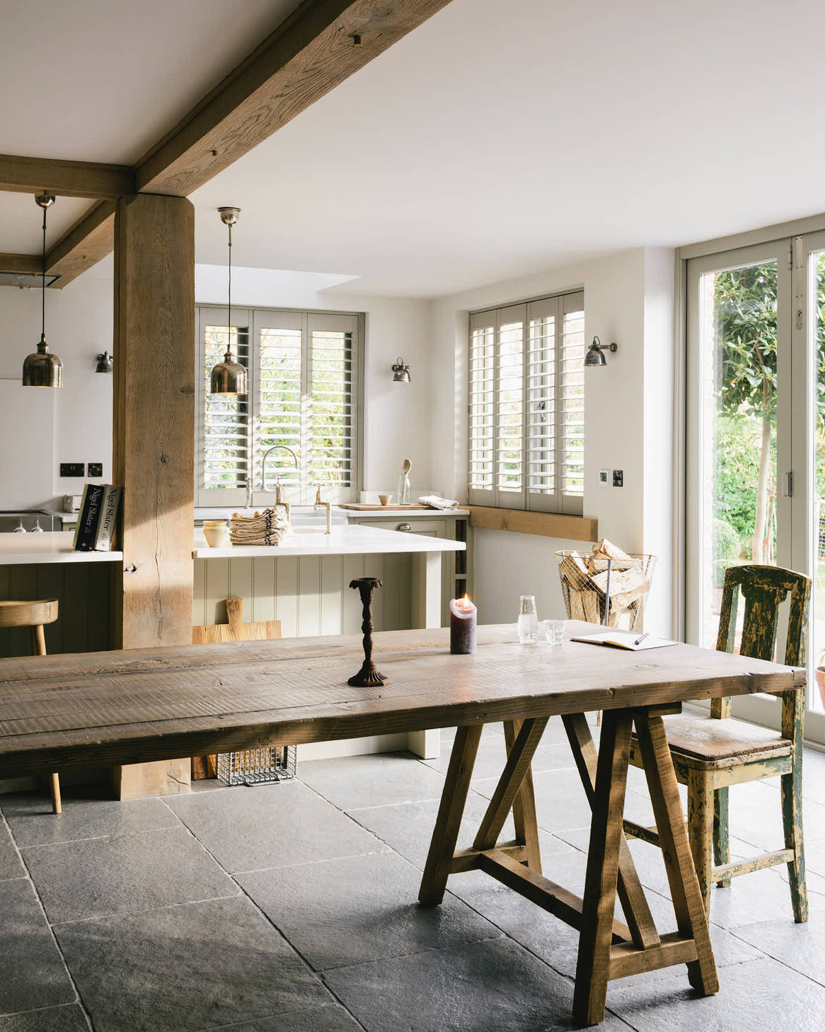 An oblique view of a tranquil and calm Shaker kitchen features a long sink and cooker run with a silestone worktop and splash back, a single Belfast sink with a nickel Ionian tap, and a long kitchen island with a breakfast nook. Beautifully decorated with a vintage dining table that matches the raw wooden beams, silver pendant lights, and a steel firewood basket, this kitchen creates a lovely, simple space that embraces natural materials.