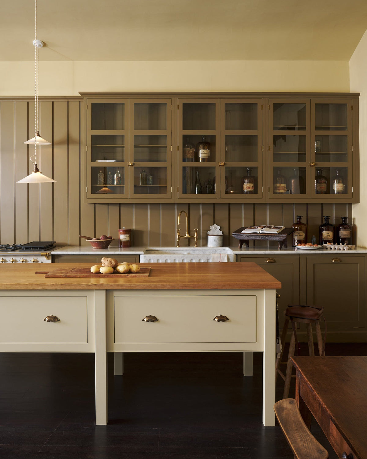 A large yet humble shaker kitchen features a long sink and cooker run with a marble worktop, a prep table with an oak worktop, a double fluted marble sink, glazed wall cupboards and a variety of antique furniture. The warm creams and greens makes the kitchen feel warm and inviting.