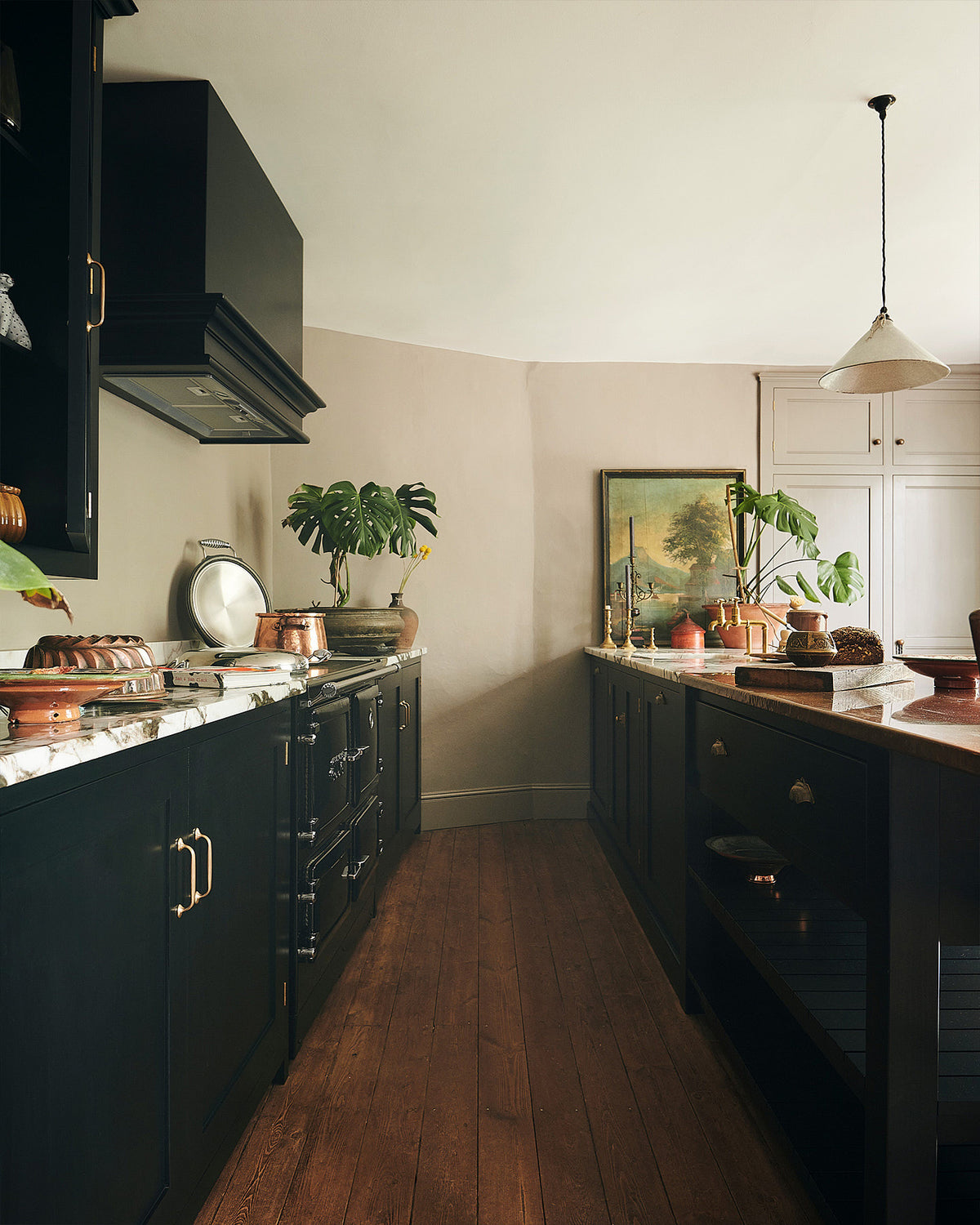 A straight view of an understated painter’s black shaker kitchen featuring an aged copper sink, aged brass taps, and a San Simone quartzite worktop—its pale white base accented with bold dark grey veins and subtle gold flecks. Grey walls frame the space, with a cheese plant and vintage oil painting adding character in the background.