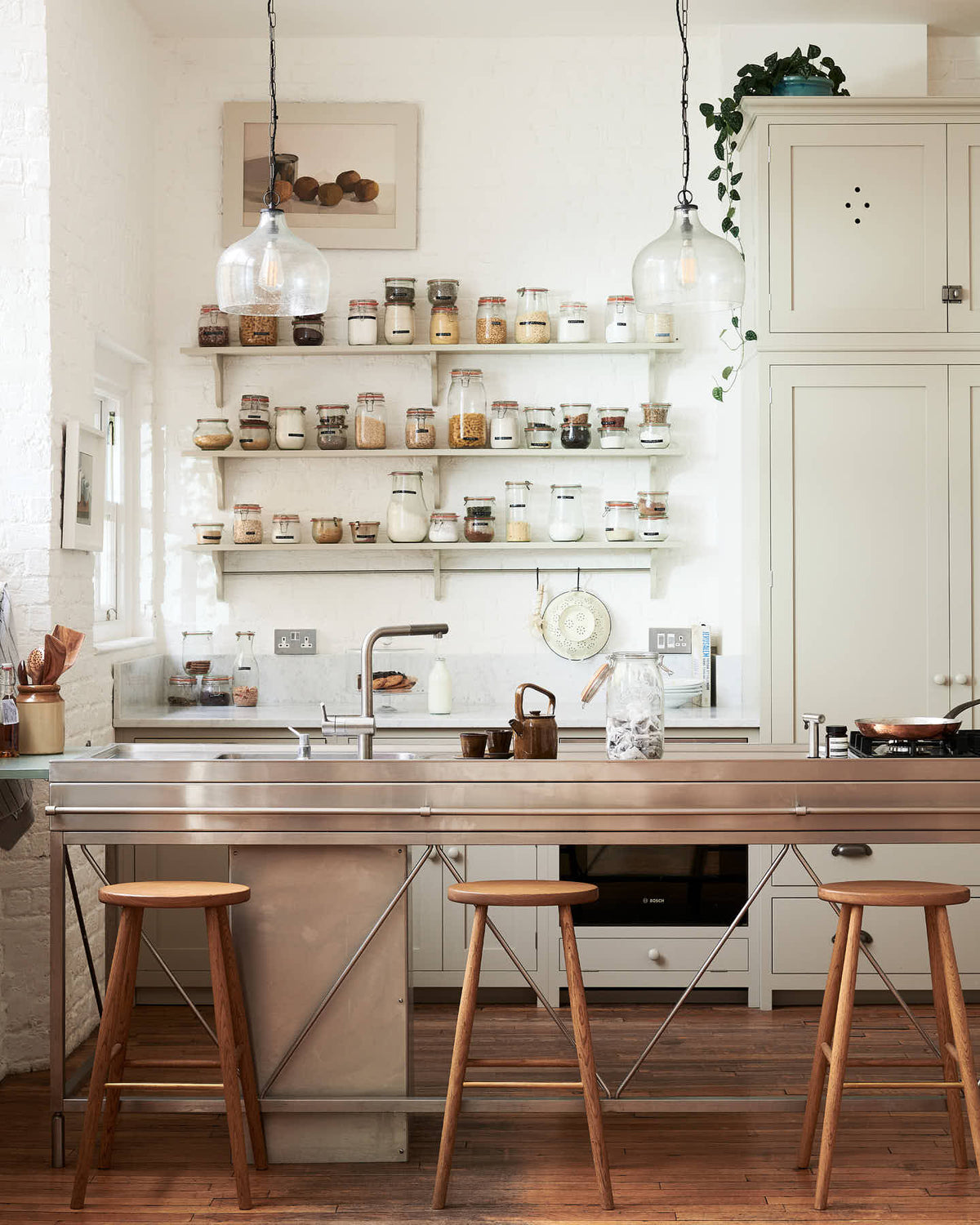 A tall and simple mushroom-coloured kitchen features a Shaker cupboard run with a full-height cabinet, a large steel cooker and sink kitchen island, and shelves with hanging rails. Adorned with Helen stools, glass jars of dried ingredients, an amber glaze teapot, glass and steel pendant lights, and still-life paintings, it creates a humble industrial kitchen space.