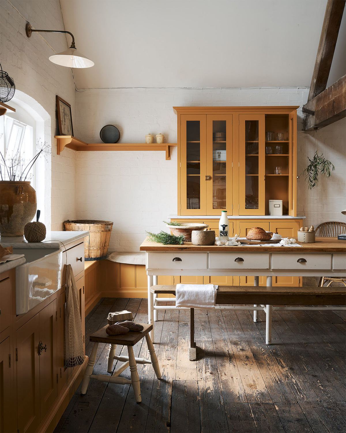 A scullery yellow shaker kitchen with a small sink run with a Carrara marble sink, a full-height cabinet, shaker shelves, an L-shaped shaker bench seat and a closed larder with glazed cupboards. The kitchen features a single Belfast sink with aged brass Mayan taps, a collection of vintage furniture and beloved items such as mixing bowls and fresh produce. The ceilings are high and retain their original exposed wooden structure.
