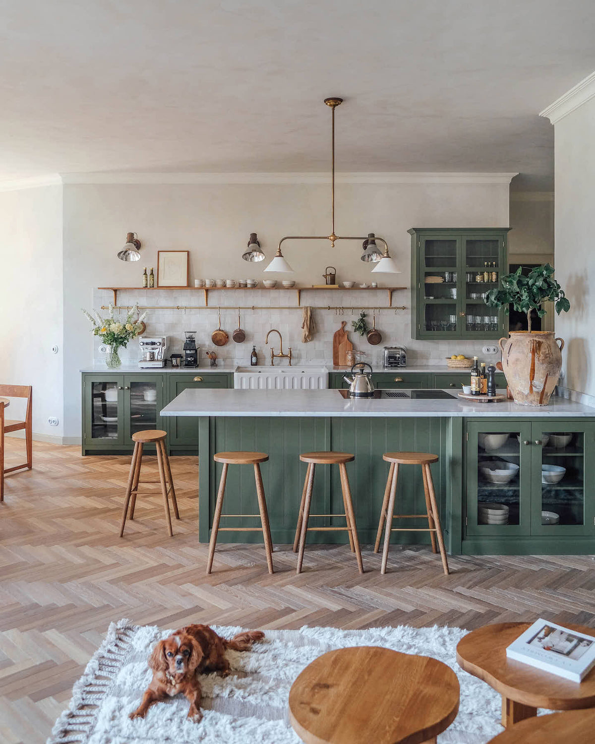 A spacious, open plan room with high ceilings, textured parquet wooden floors and a beautiful ‘Wilkes Green’ painted Shaker kitchen. The walls are painted in a creamy, lime wash paint and the backsplash is fitted with glossy beige tiles. A large kitchen island dominates the image, with four Helen’s Stools pulled up to it. The room feels fresh and contemporary but still has lots of traditional charm.