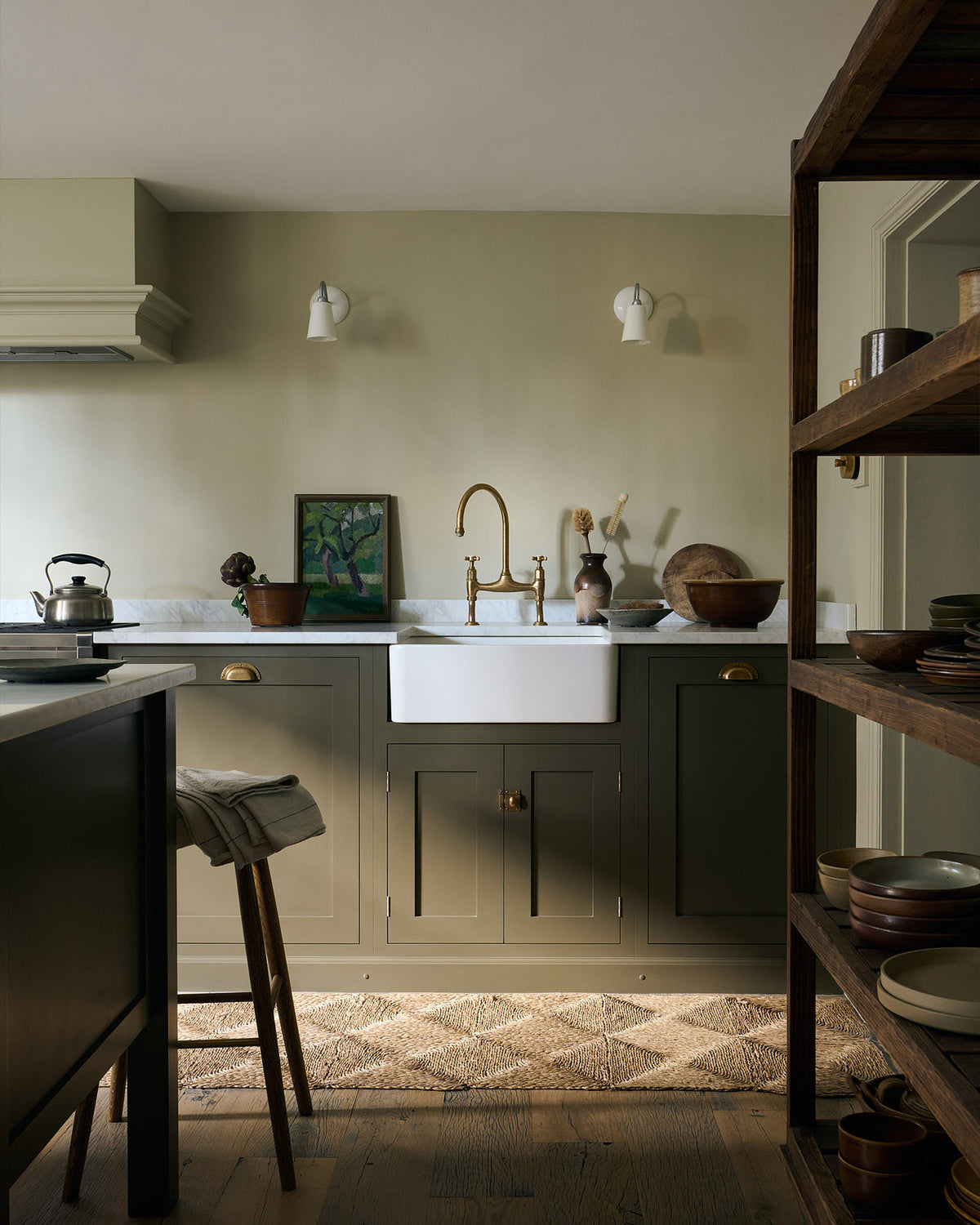 A closer look at an olive green Shaker kitchen featuring a sink and cooker run, a freestanding prep table with a Carrara marble worktop, and Helen Stools gathered around the breakfast nook. A large wooden shelf displays a thoughtful selection of muted ceramics and mugs. The space embraces a simple theme rooted in raw, natural materials—an ode to Mother Nature—highlighted by a small oil painting of trees placed beside the single Belfast sink with an aged brass Ionian tap.