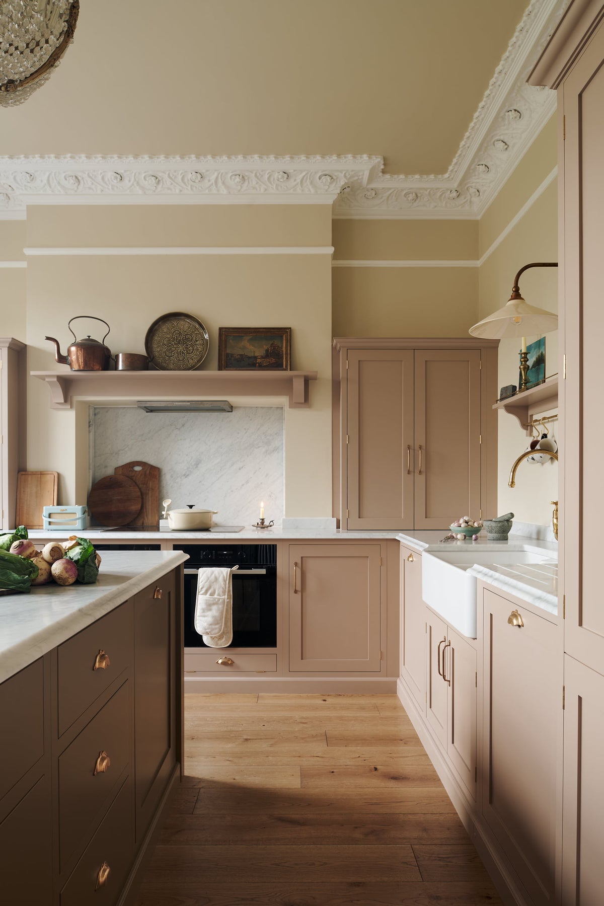 An elegant and soft Princelet pink Shaker kitchen unfolds with an L-shaped cooker and sink run topped in Carrara marble. Alongside countertop cupboards, a kitchen island with a breakfast nook, and an over mantle above the alcove of a chimney breast and marble splash back, it create a beautifully functional space.