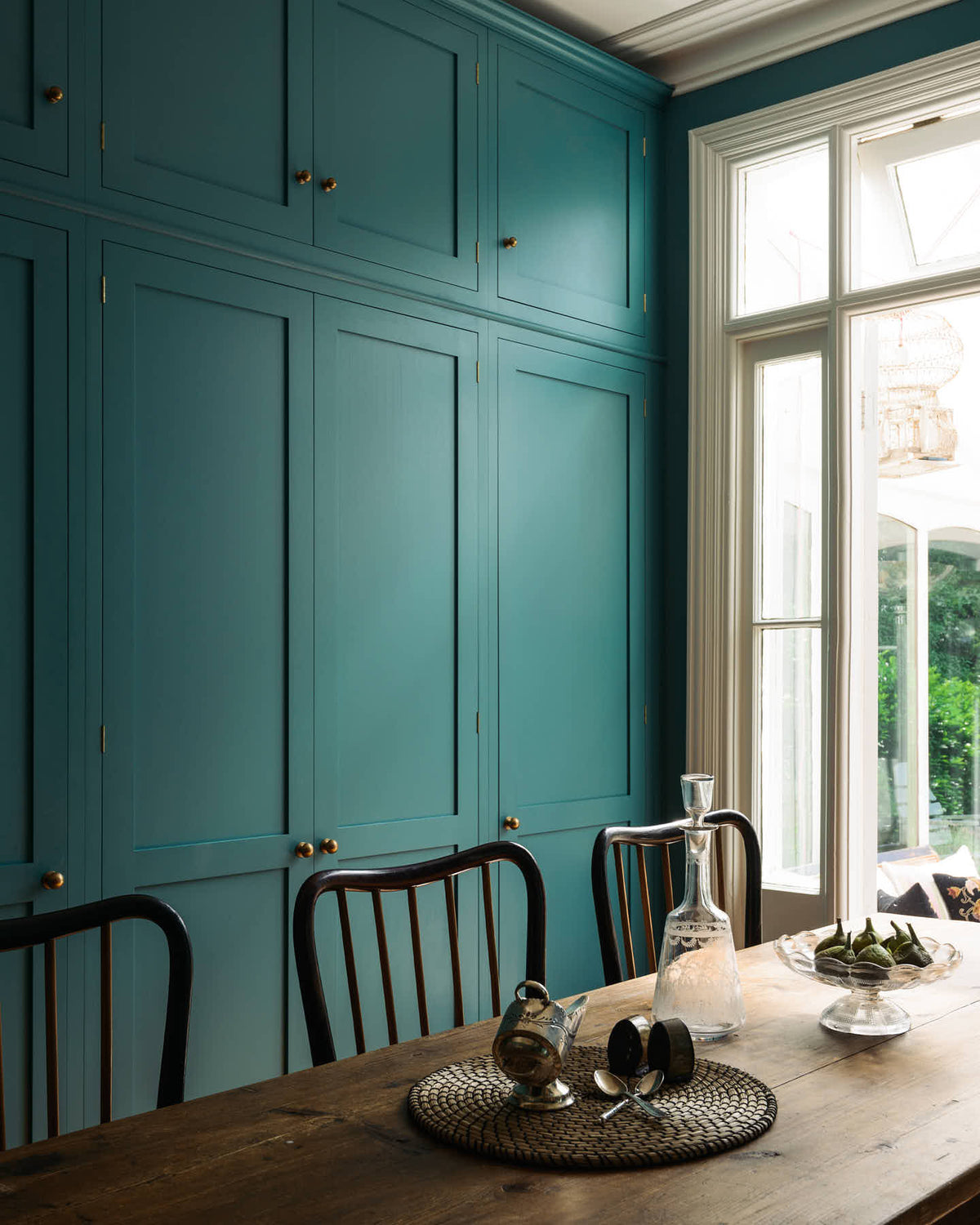 An angled view reveals a grand, sea foam green Shaker kitchen featuring a bank of full-height pantry cupboards with a Carrara marble worktop, and a charming French door leading to the conservatoire. Adorned with beautiful vintage furniture, a glass bowl of green figs, antique silverware, and a decanter, the kitchen reflects the refined tastes of its socialite and collector owners.