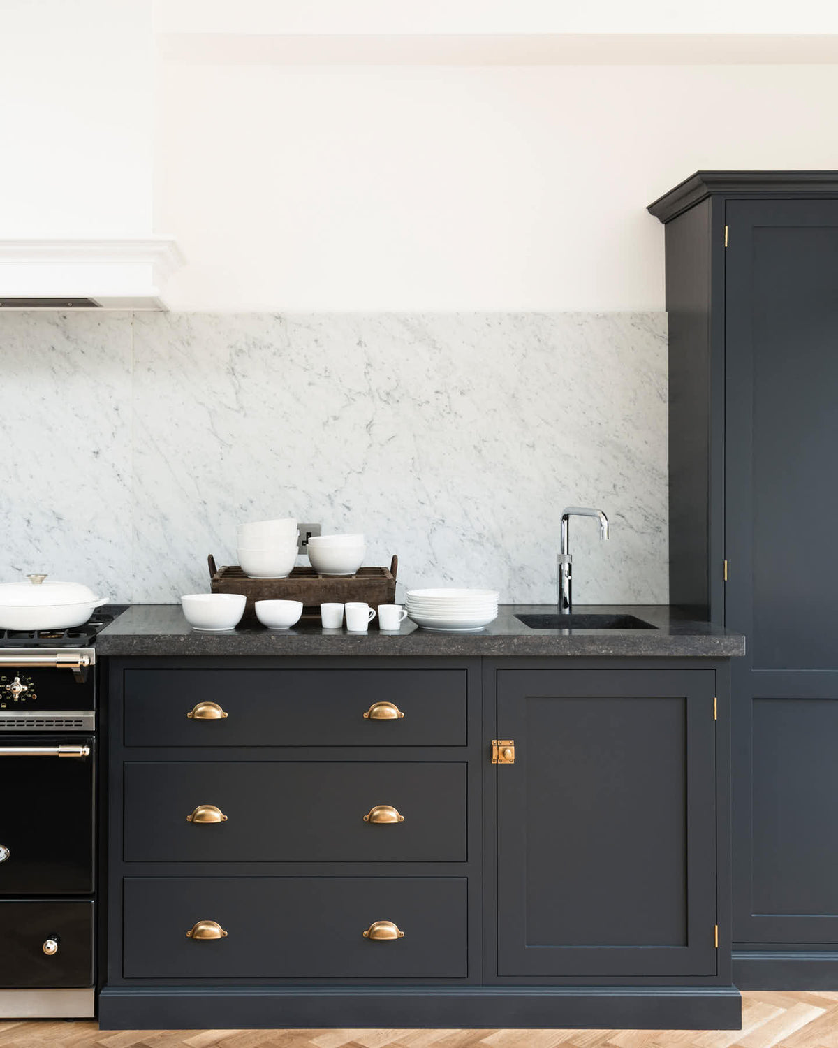 A corner of an oblique view of a bright and sophisticated pantry-blue Shaker kitchen features a sink and cooker run with a Belgian blue limestone worktop, a Carrara marble splash back, and full-height cupboards.  Adorned with a silver tap, a single copper sink, a cactus plant, and beautiful brass fittings, it creates a functional and contemporary kitchen space.