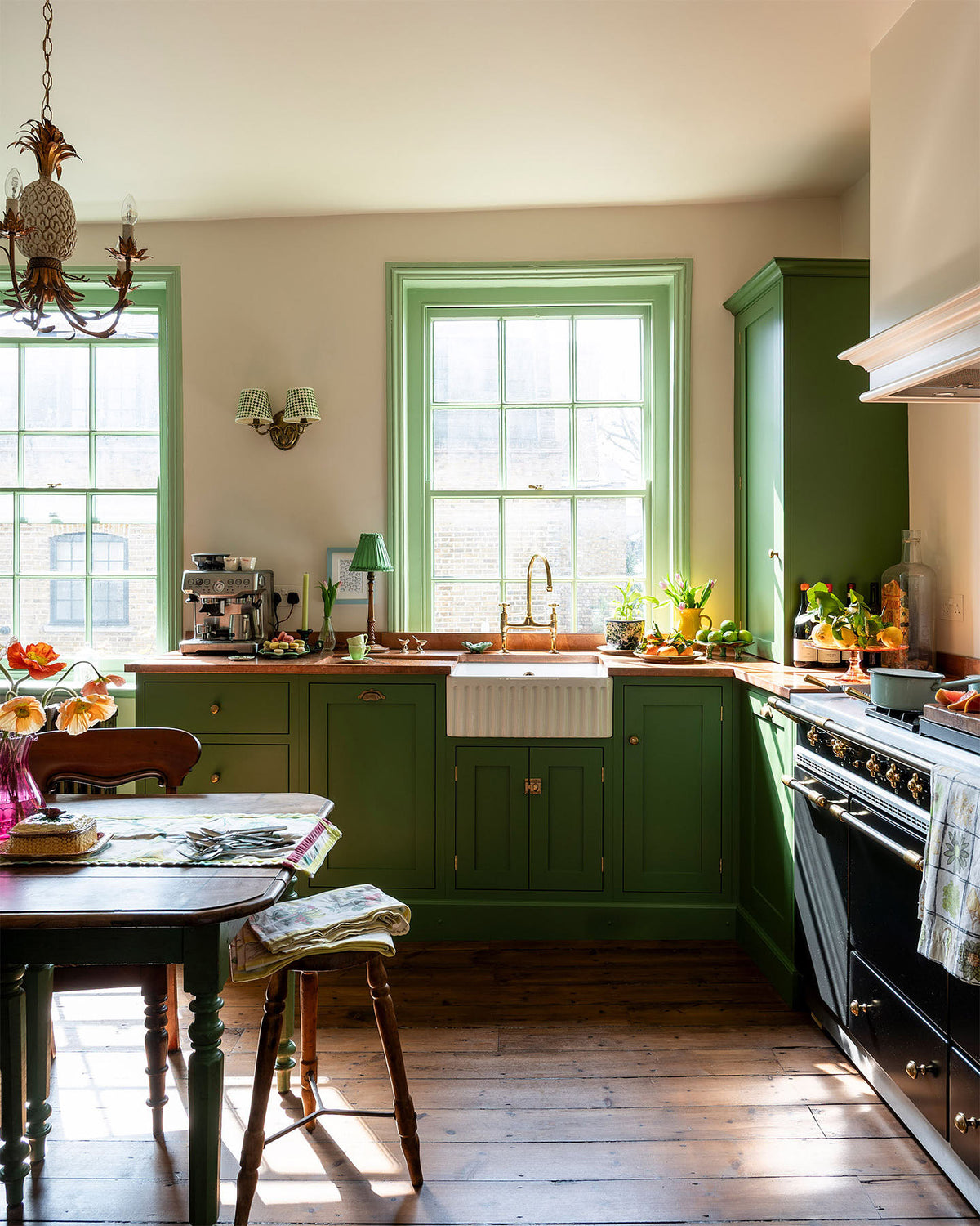 A bright and humble lime-green kitchen with an L-shaped cooker and sink run topped with aged copper worktops, a countertop and glazed cupboards, a single fluted Belfast sink with an aged brass Ionian tap, and colourful accents like tulips, a bowl of limes, macaroons, and a golden pineapple chandelier.