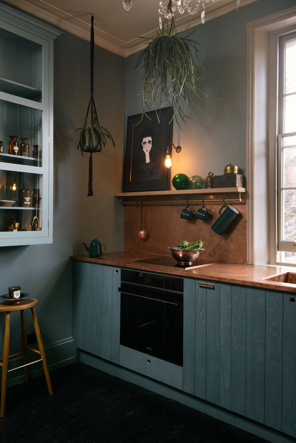 An angled view of a sweet and simple Clerkenwell blue Sebastian Cox kitchen features a short cooker and sink run with a copper worktop and splashback, a single copper sink, an electric stove, shelves with brass hanging rails, and a glazed wall cupboard. Adorned with artisan ceramics, vintage bric-a-brac, hanging plants, a sweetheart cabbage, a framed quirky illustration, and a bare wall light, this kitchen creates a glamorous and practical space.