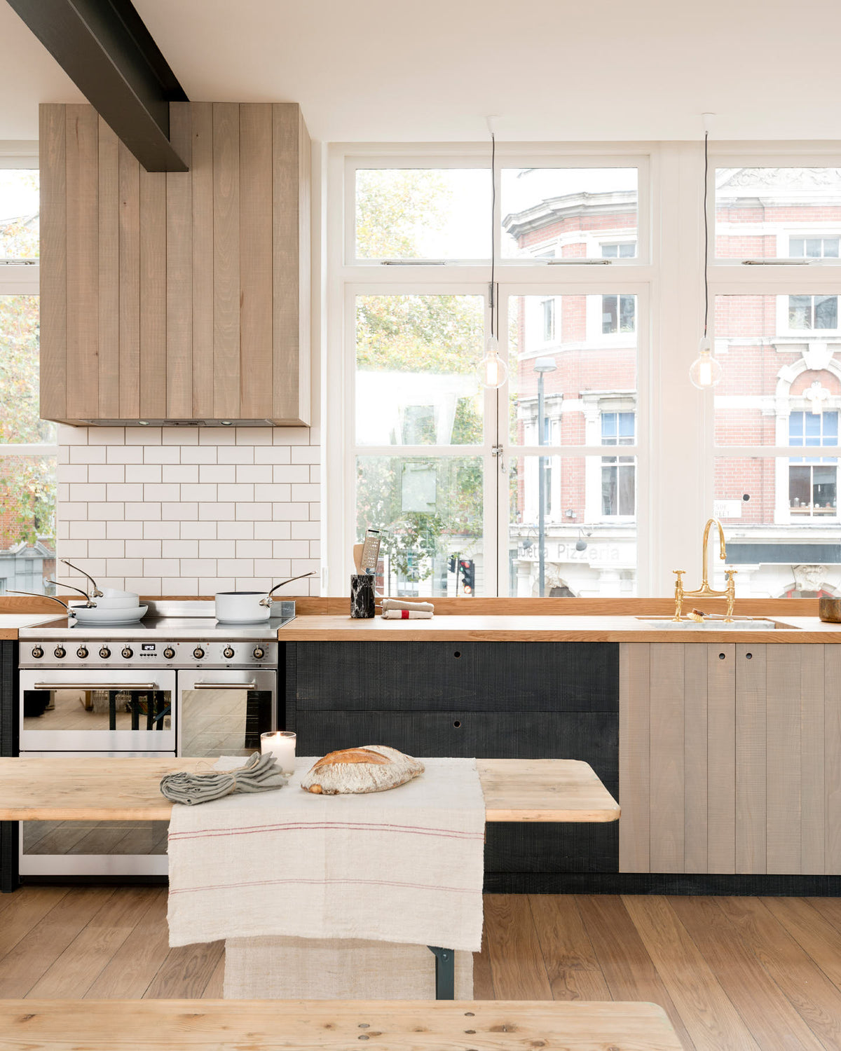 A simple and contemporary Sebastian Cox kitchen features a long sink and cooker run with a solid oak worktop, a single Belfast sink, an aged brass Ionian tap, a canopy, and a silver electric stove. Adorned with bare pendant lights, an Oktoberfest bench and table, white pans, a loaf of bread, and a natural linen table runner, this kitchen creates a cosy space with industrial details and a practical finish.