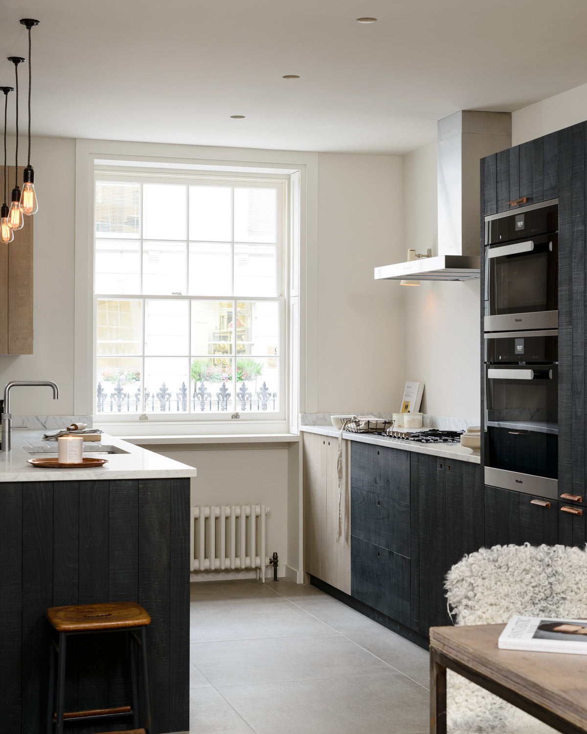 An oblique view of a sleek and modern Sebastian Cox galley kitchen features a sink and cooker run with Carrara marble worktop, a transom partition window, a glazed wall cupboard, an appliance cabinet, and a fridge/freezer cupboard. Adorned with simple ceramics, bare pendant lights, a wooden dining table and chairs, and a fluffy wool blanket, this kitchen creates an elegant and practical space.