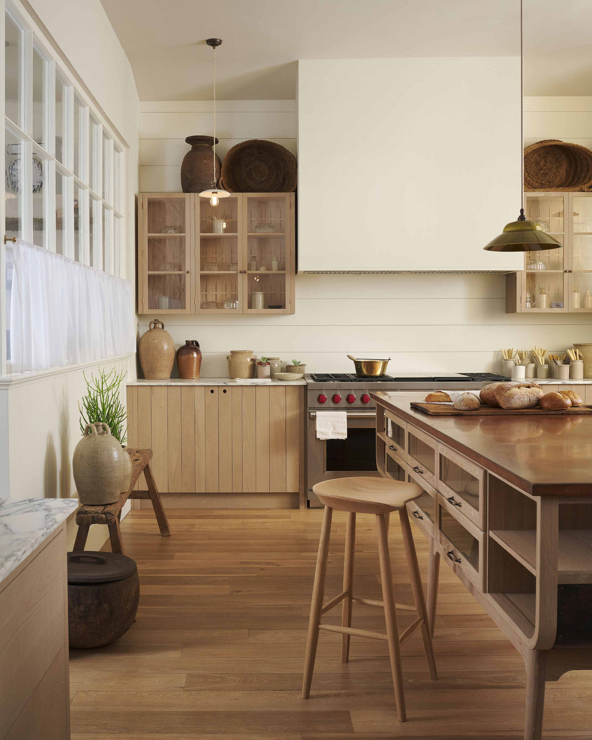 An all-natural and functional Sebastian Cox kitchen features a cooker and sink run with a marble worktop, glazed wall cupboards, and an iconic Haberdasher’s prep table with a copper worktop. Lovingly adorned with antique artisan ceramics, woven baskets, beautiful houseplants, and beloved figurines, it creates a wonderfully simple kitchen with a love of natural sustainability. 
