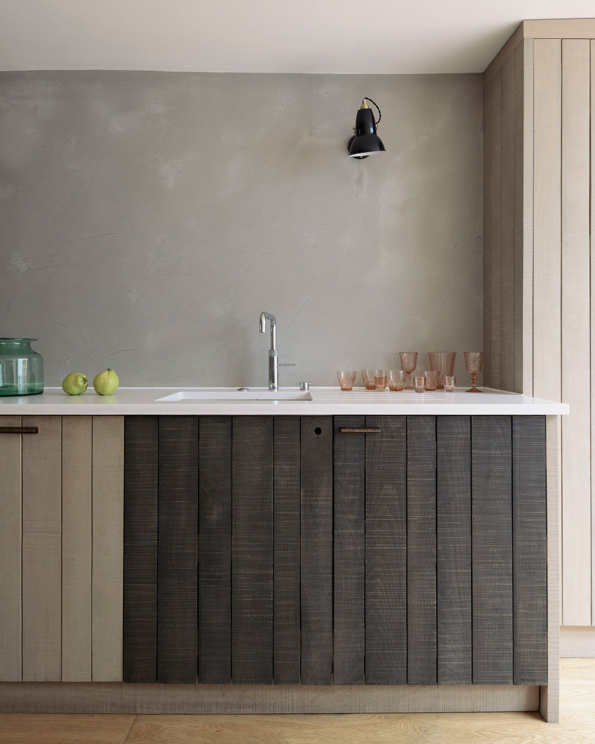 A close-up of a sleek and simple Sebastian Cox kitchen includes a cooker and sink run topped with a silestone worktop, a full-height larder, a single Belfast sink, a silver tap, and walls finished with natural limestone. The space is beautifully decorated with red-tinted drinking glasses and a black industrial wall lamp, creating a minimal yet earth-conscious and sustainable kitchen environment.