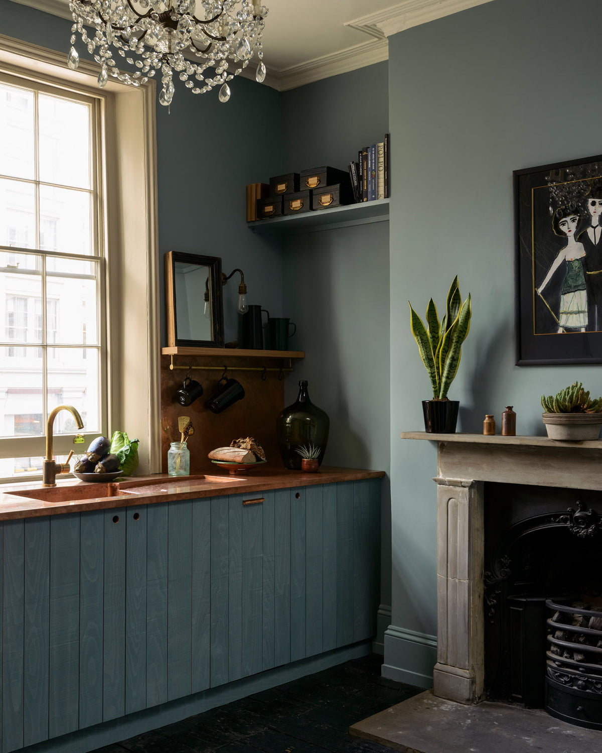 An angled view of a sweet and simple Clerkenwell blue Sebastian Cox kitchen features a short cooker and sink run with a copper worktop and splash back, a single copper sink, an electric stove and shelves with brass hanging rails. Adorned with artisan ceramics, vintage bric-a-brac, aubergines, hanging plants, a framed quirky illustration, a crystal chandelier, and bare wall lights, this kitchen creates a glamorous and practical space.