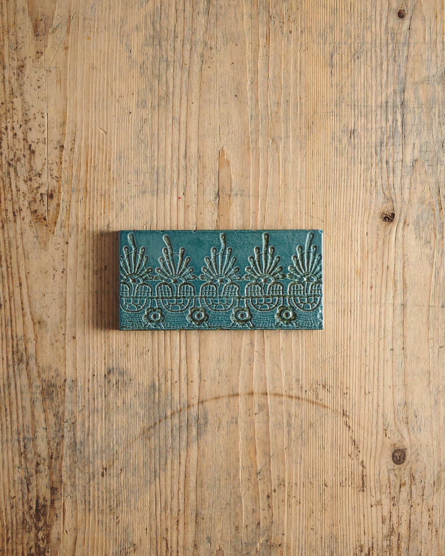 A glossy teal metro tile with a decorative lace pattern on a rustic wooden surface.