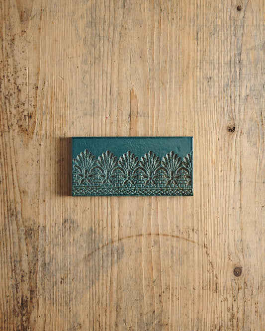 A glossy teal metro tile with a decorative lace pattern on a rustic wooden surface. | Flemish Metro