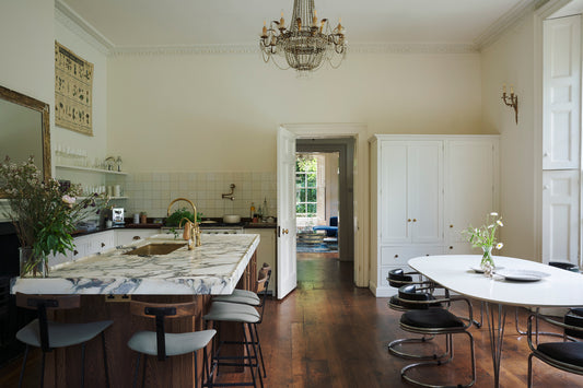 A bright, elegant kitchen and dining area with a white marble island, dark wood floors, a white dining table with black chairs, and a large white cabinet.