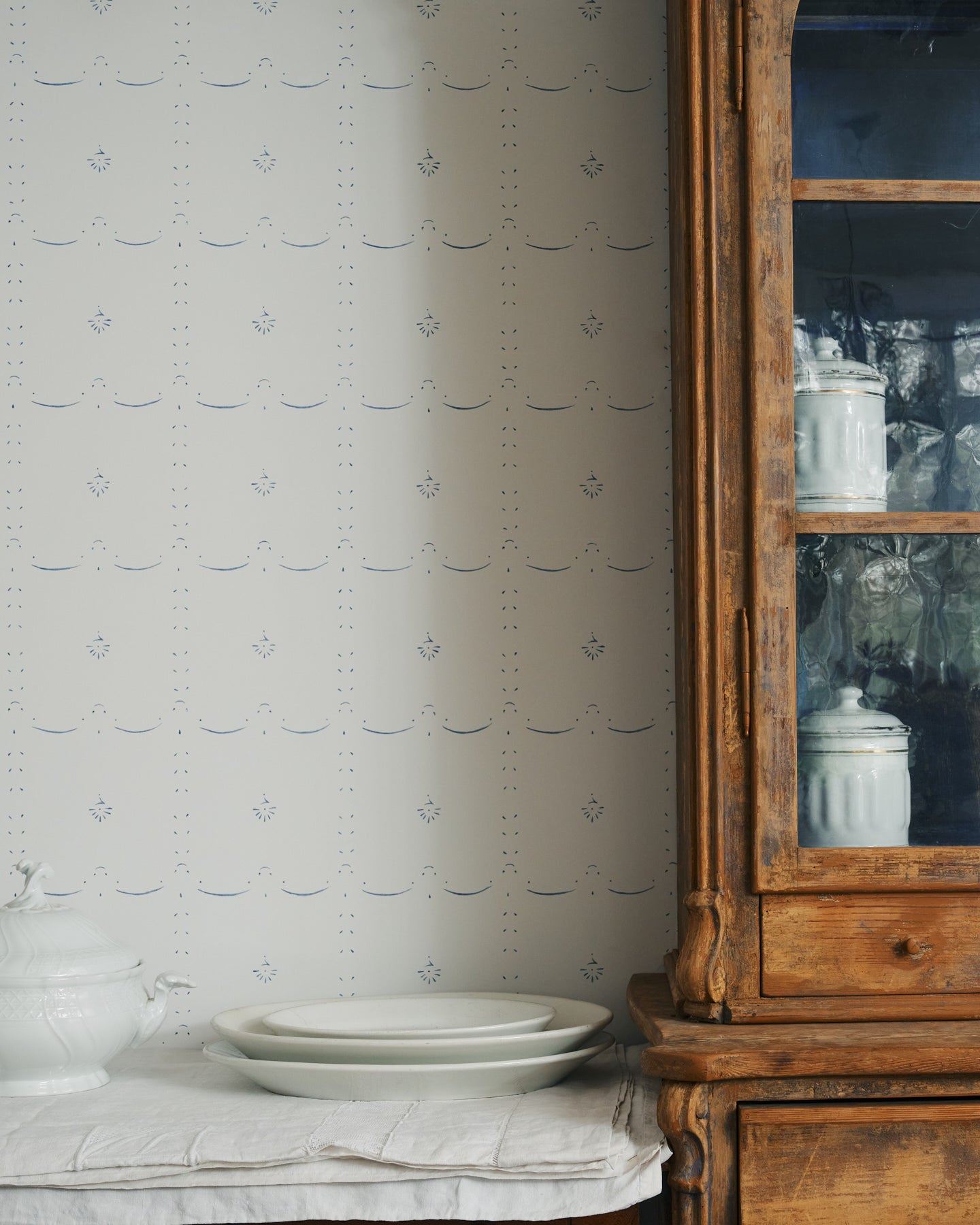 An off-white wallpaper with a delicate hand-painted pattern in a blue colour around a vintage cupboard and console table.