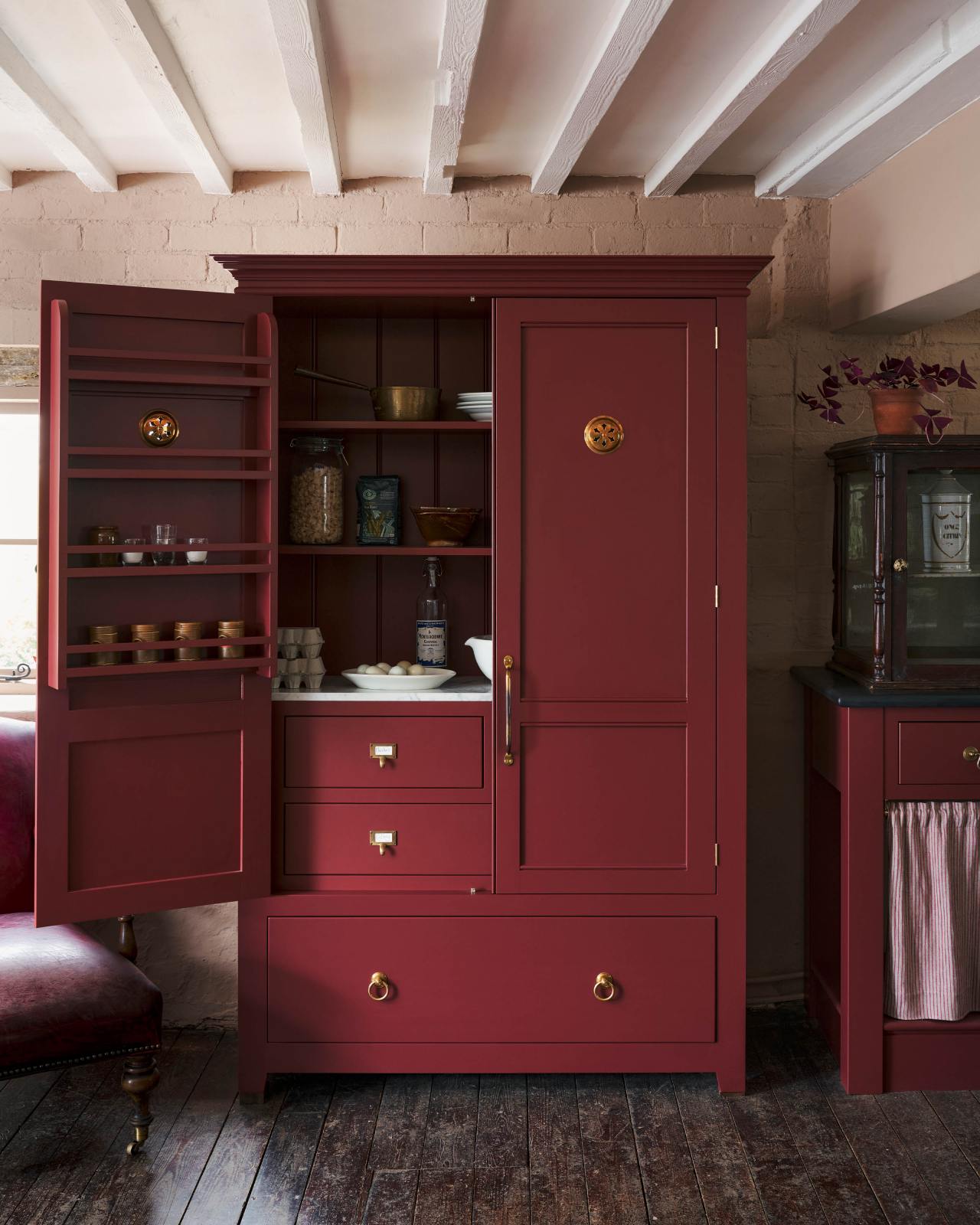 A freestanding pantry painted in a deep red hue is fitted with brass details such as ventilation grilles, hinges, ring pulls an external drawer and cardframe pulls on internal drawers. The room is rustic with scuffed wooden floorboards, painted brick walls and painted white beams overhead.