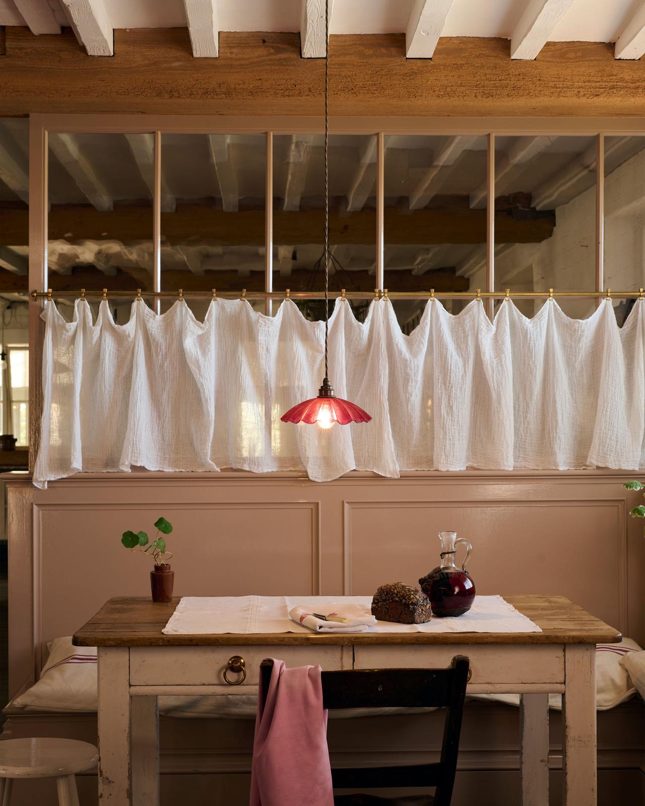 A small kitchen table with bench seating behind and a pretty white curtain above it. There is a low hanging pink pendant over the table, it is made from glass that has been formed into pretty pleats.