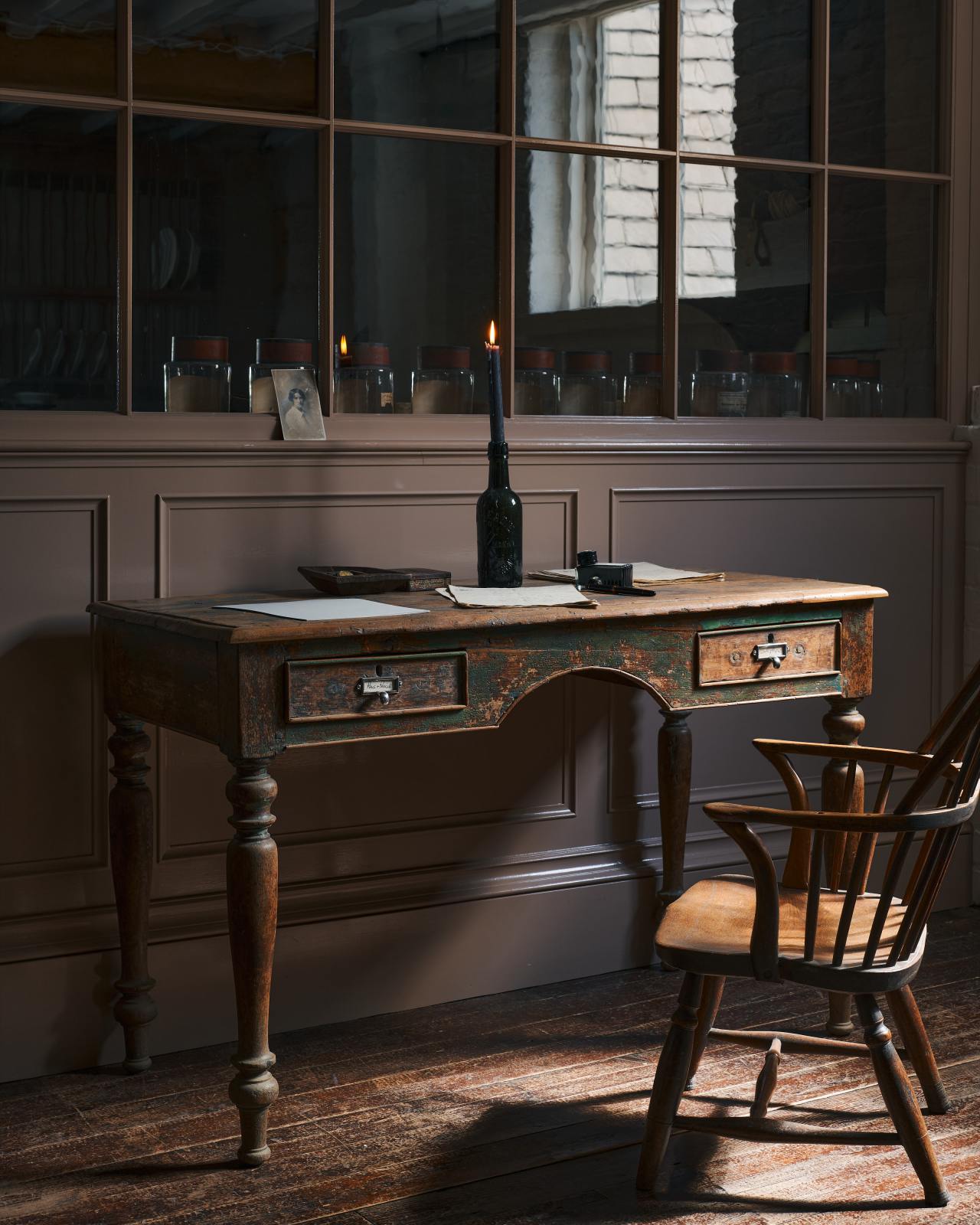 Antique silver pulls are attached to a vintage desk with chippy green paint so much of the original timber shows through. On the desk is a dark glass bottle with a lit candle, and various vintage papers, as well as an ink pen and ink bottle. The pulls themselves are a dark silver in colour, each with a paper insert describing the contents of the drawers, 'pens and pencils' and 'writing paper'.