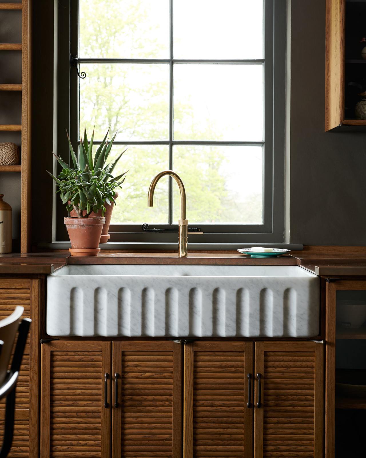 A front of shot of deVOL's Haberdasher's Kitchen at their Cotes Mill showroom. In the centre of the image is a sink cabinet with tambour-fronted doors below, housing a 1000mm double bowl ceramic sink with a fluted front. The tap is brass and the worktops around the sink is aged copper. There is a window behind the sink and dark grey walls either side.