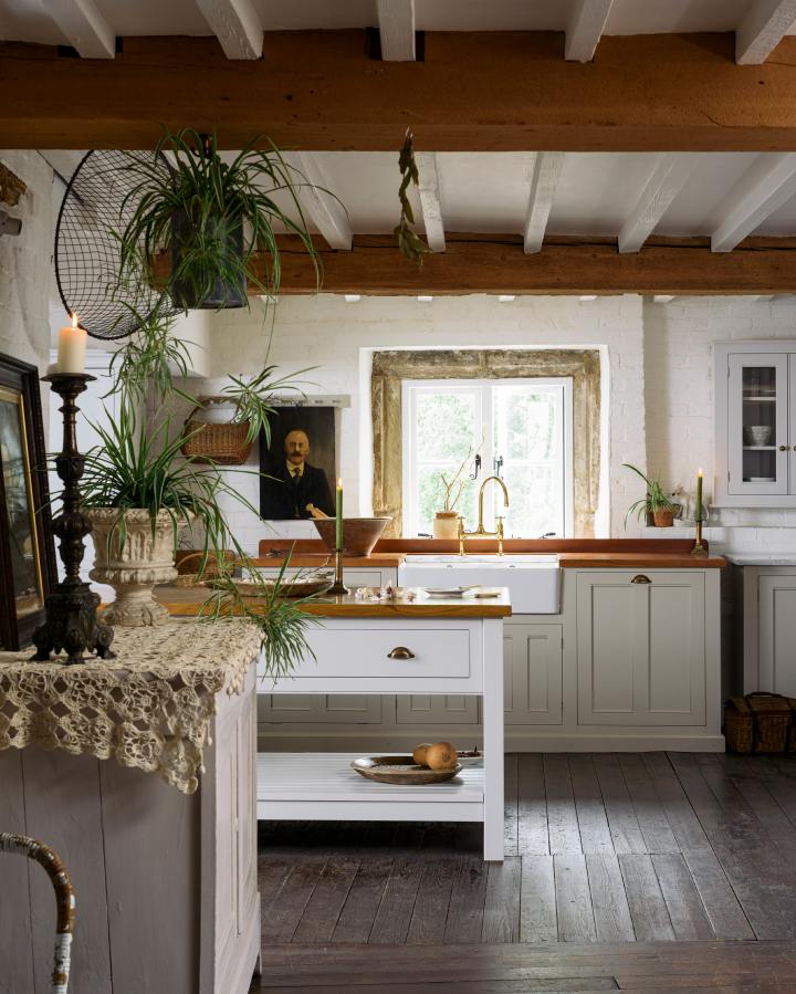 A room with low beamed ceilings and a kitchen made up of freestanding pieces of furniture by deVOL. The floors are dark original wooden planks, the cupboards are painted in two pale and fresh shades and the walls are white. A prep table and sink cabinet are topped with warm wooden worktops and there is a very old window looking out onto lush grounds beyond.