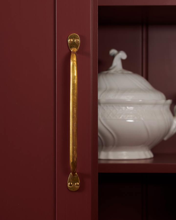 A close-up detailed shot of a large deVOL ‘Pull Handle’, fitted to the door of a maroon-coloured pantry cupboard. The golden ‘Heirloom Brass’ finish reflects the warm tones of the cupboard to create a subtle glow.