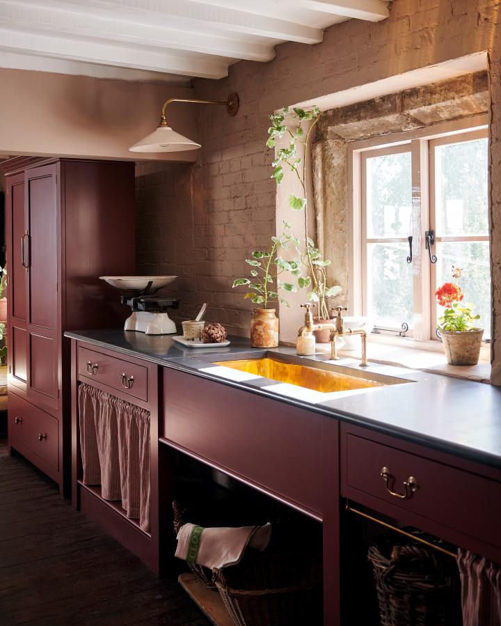 An oblique view of a Victorian-style country house sink cabinet, painted in a dark maroon colour. Strong warm light steams through a window and reflects dramatically off aged brass bibcock taps and a polished undermounted brass sink. The glamour is contrasted by the honed slate worktop and the rustic styling, with large wicker baskets, worn ceramic pots and climbing geraniums.