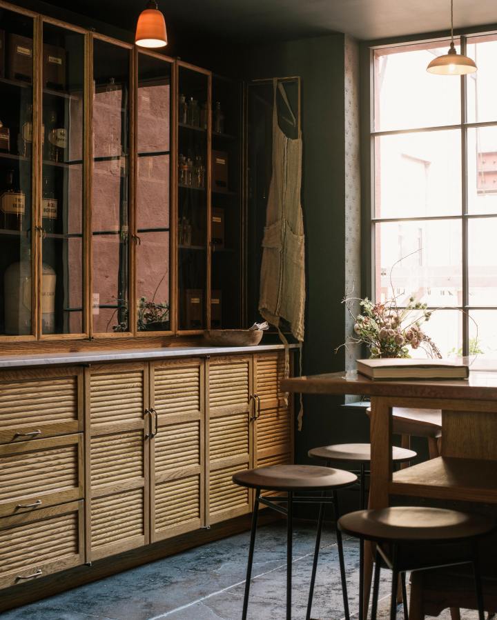 A dark room with charcoal-coloured walls and ceiling and a riven black stone floor, contrasting with the amber-coloured mid-century-style oak kitchen furniture. In the foreground, there are two industrial-style metal framed stools and a prep table with turned oak legs, an open curved end, glazed drawers full of rustic pottery and a mottled copper worktop. Behind is a tall bank of glazed cupboards full of antique chemists' jars and tins, each with a handwritten label.