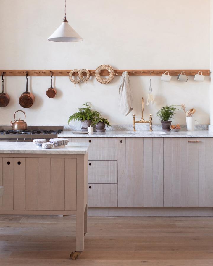 A light contemporary kitchen with creamy textured walls, rough-sawn beech slatted cupboard doors, an aged brass tap and a span of wooden pegs for hanging mugs, pans and utensils above a mottled white marble worktop. The styling is simple, Scandinavian and slightly rustic, with small potted ferns and woven rope trivets. 