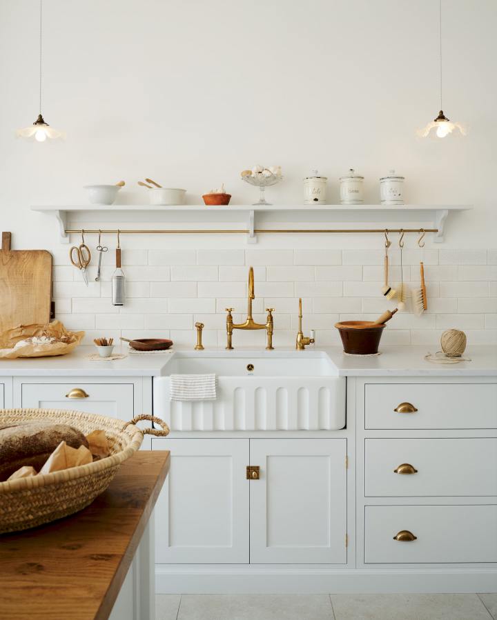 A straight on shot of a shaker-style kitchen sink run. The cupboards have been painted in deVOL's Shaker colour, 'Damask'. It's a very pale off white with a hint of grey. The sink is a ceramic fluted style and there are two brass taps and a rinse installed. The backsplash is made up of metro tiles and there is an open shelf above with a hanging rail.