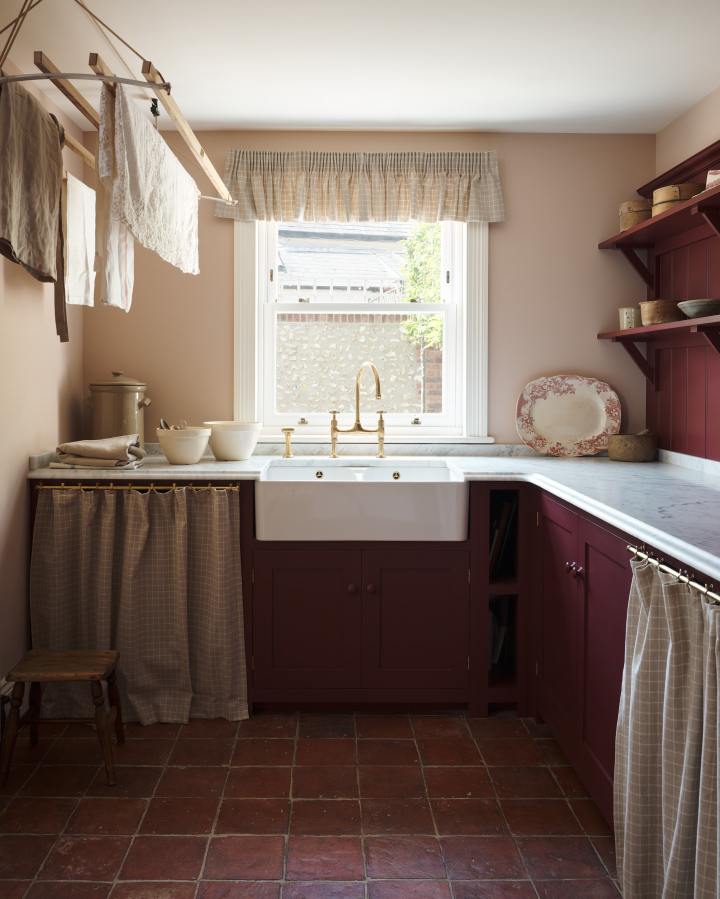 An ‘L’ shaped laundry room with fitted Shaker base cabinets painted in a dark, berry-red shade and linen curtains hanging from aged brass rails to hide the appliances. A wooden laundry maid and rustic terracotta tiled floor give the space a utilitarian feel, whilst delicate pink walls and honed marble worktops add a little bit of luxury.