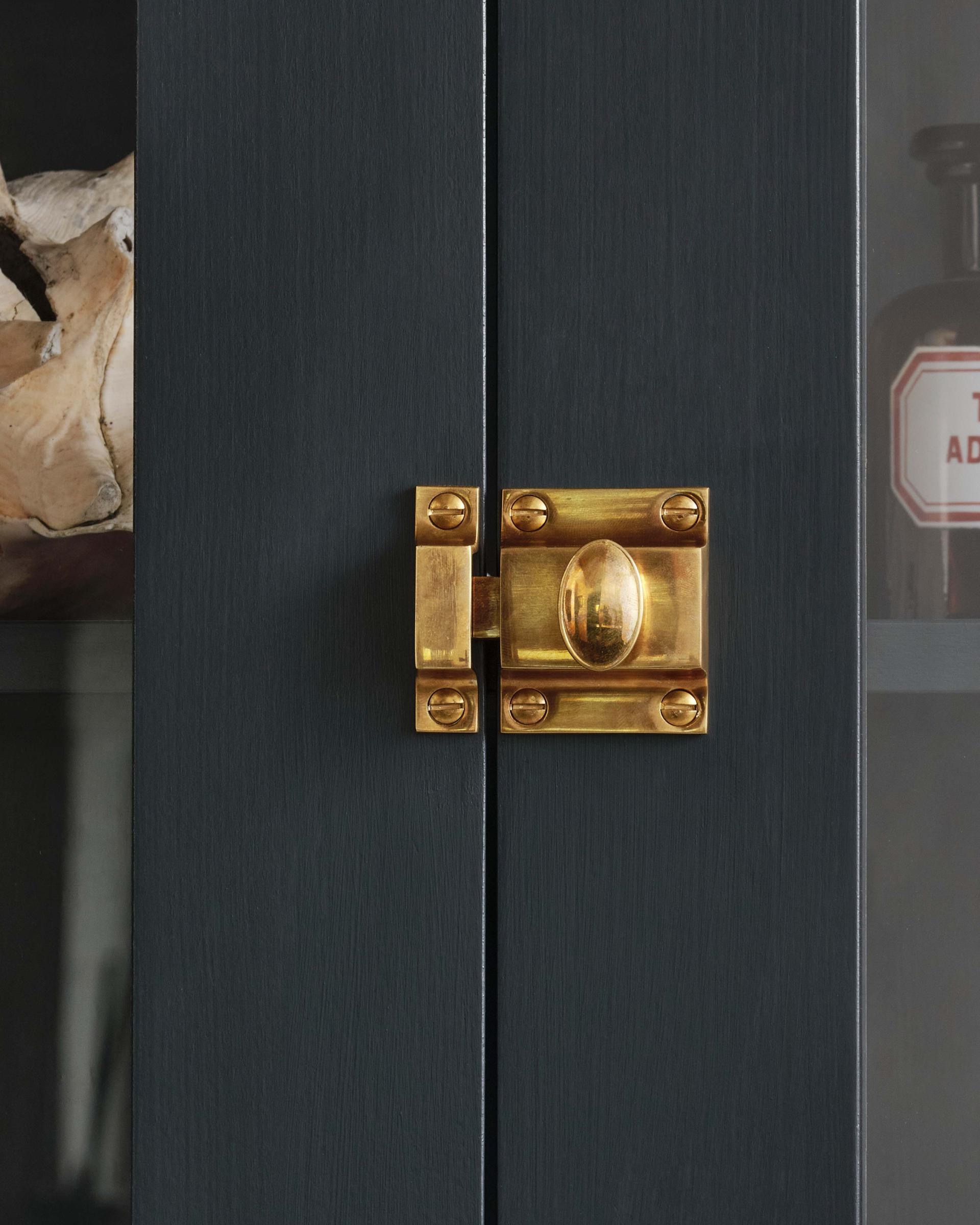 Classic brass cabinet catch with an aged, heirloom finish, featuring a traditional design and mounted on a light wooden door.