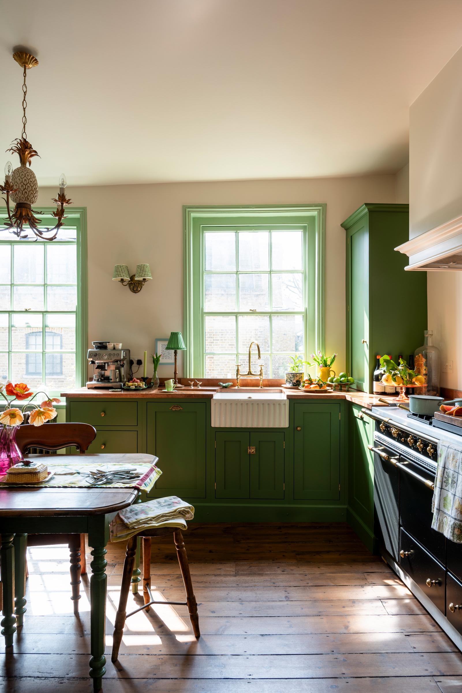 A cozy view of a classic kitchen featuring deep green shaker cabinets, copper counters, and sash windows. The ceramic sink and potted plants bring warmth, with a glimpse of the stovetop and coffee station in the background