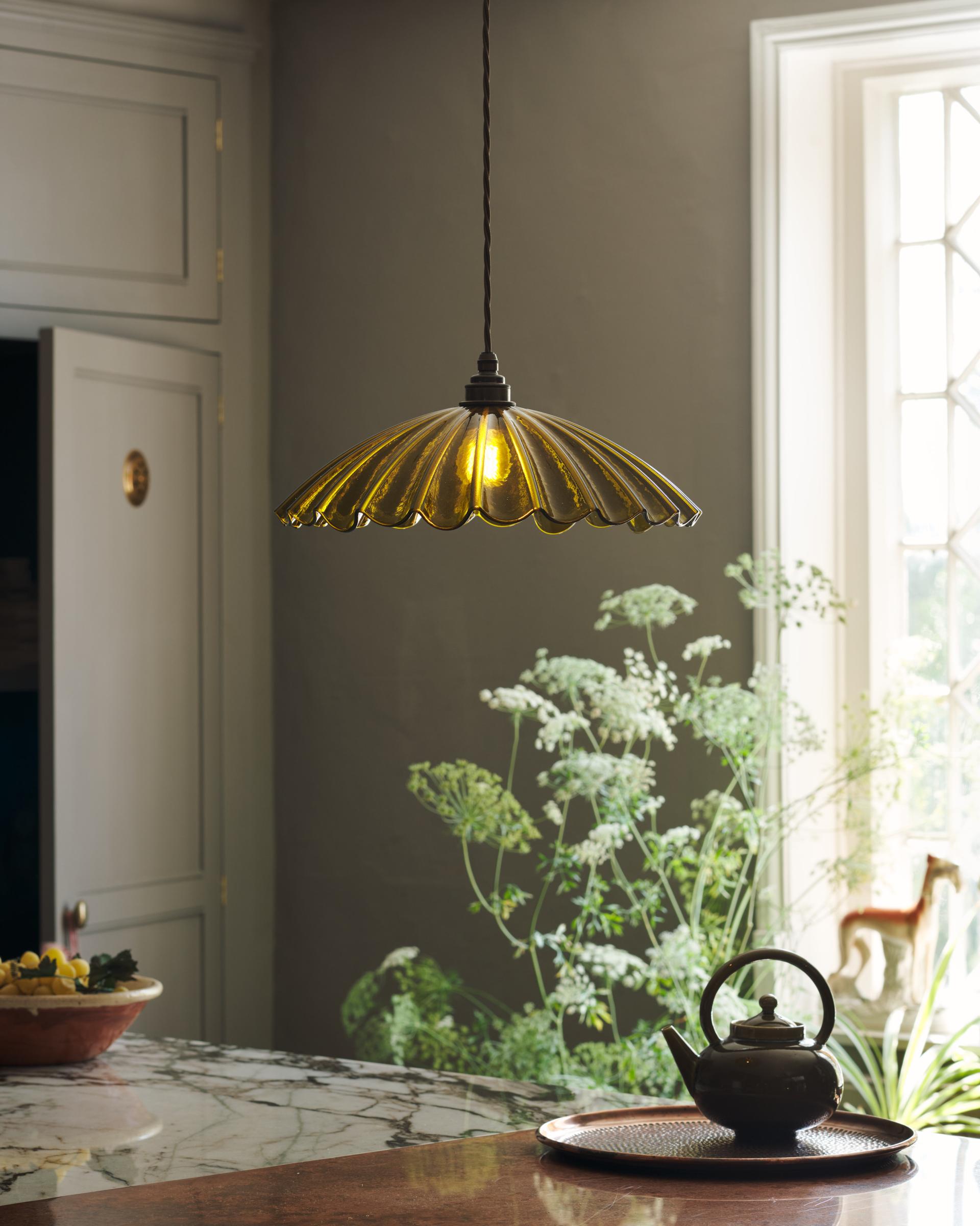 A close-up shot of a delicate frilly glass pendant light in a unique chartreuse colour fitted above a copper and marble kitchen island. The light creates a golden glow through the green-coloured glass, picking up on the hints of copper and aged brass throughout the room.