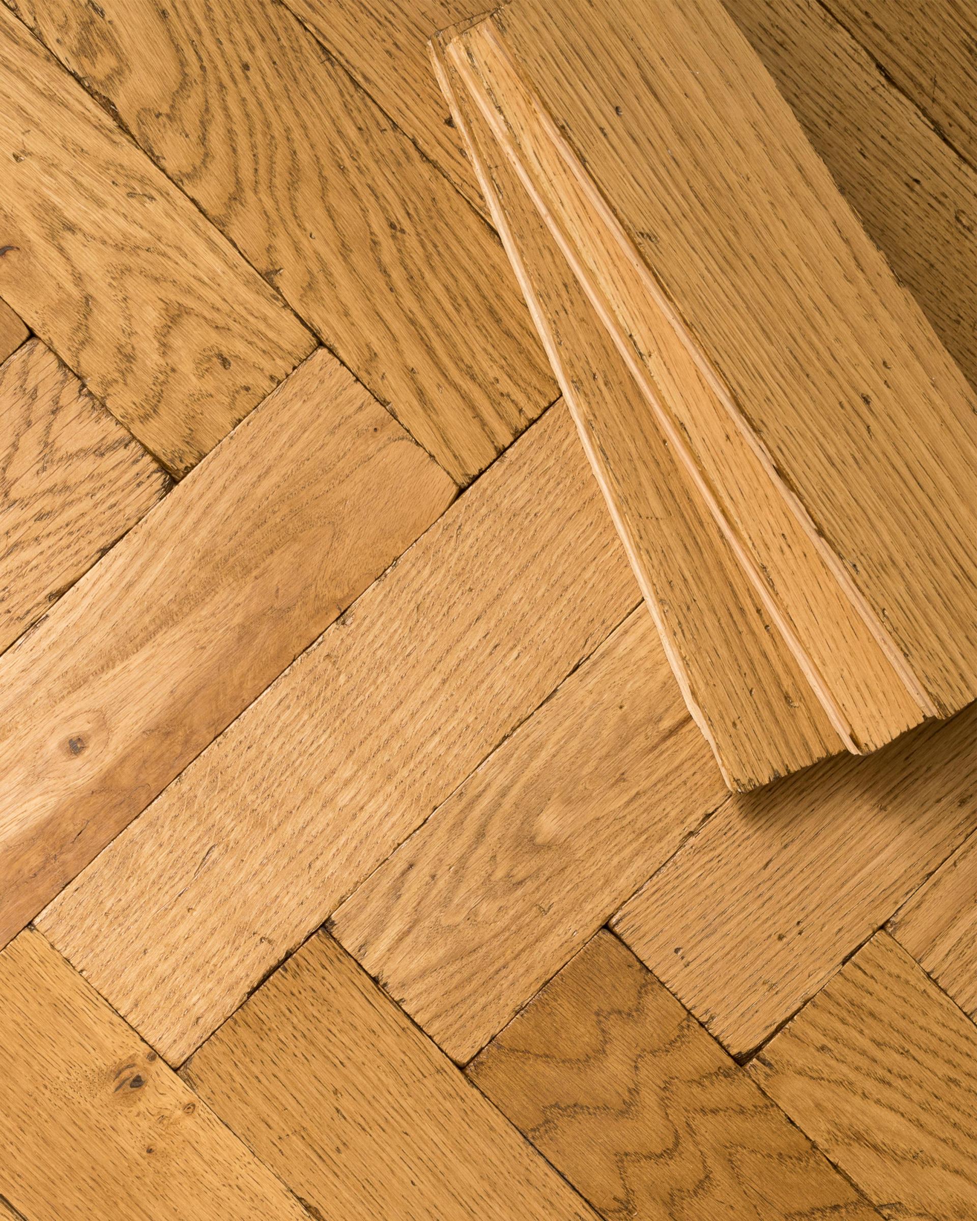 A close-up overhead view of oak parquet flooring with a warm aged finish. Three loose blocks are stacked in one corner.
