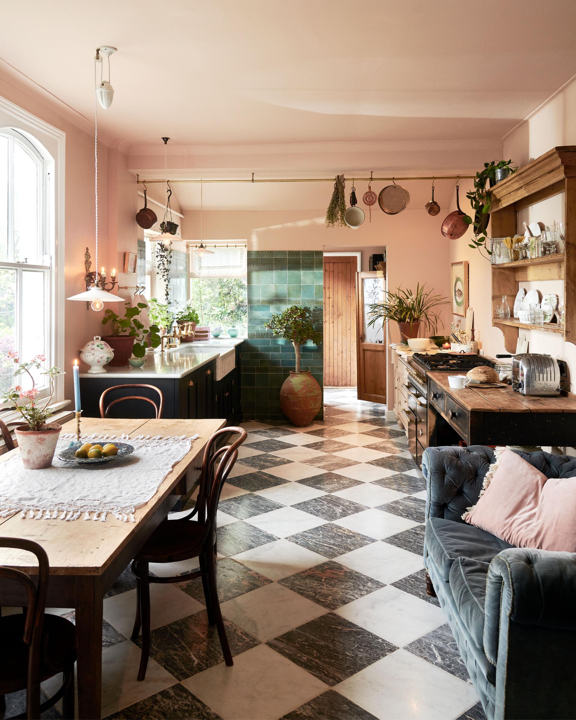 A bright peach-coloured room with a chequerboard marble floor and an eclectic collection of vintage furniture, including a table and chairs, a prep table and a decorative set of drawers. In the left corner is a modest sink run with black painted Classic English cupboards, a Carrara marble worktop and brass fittings. Pots and pans hang from a brass ceiling rail, mixed with a collection of vintage milk glass pendant lights.