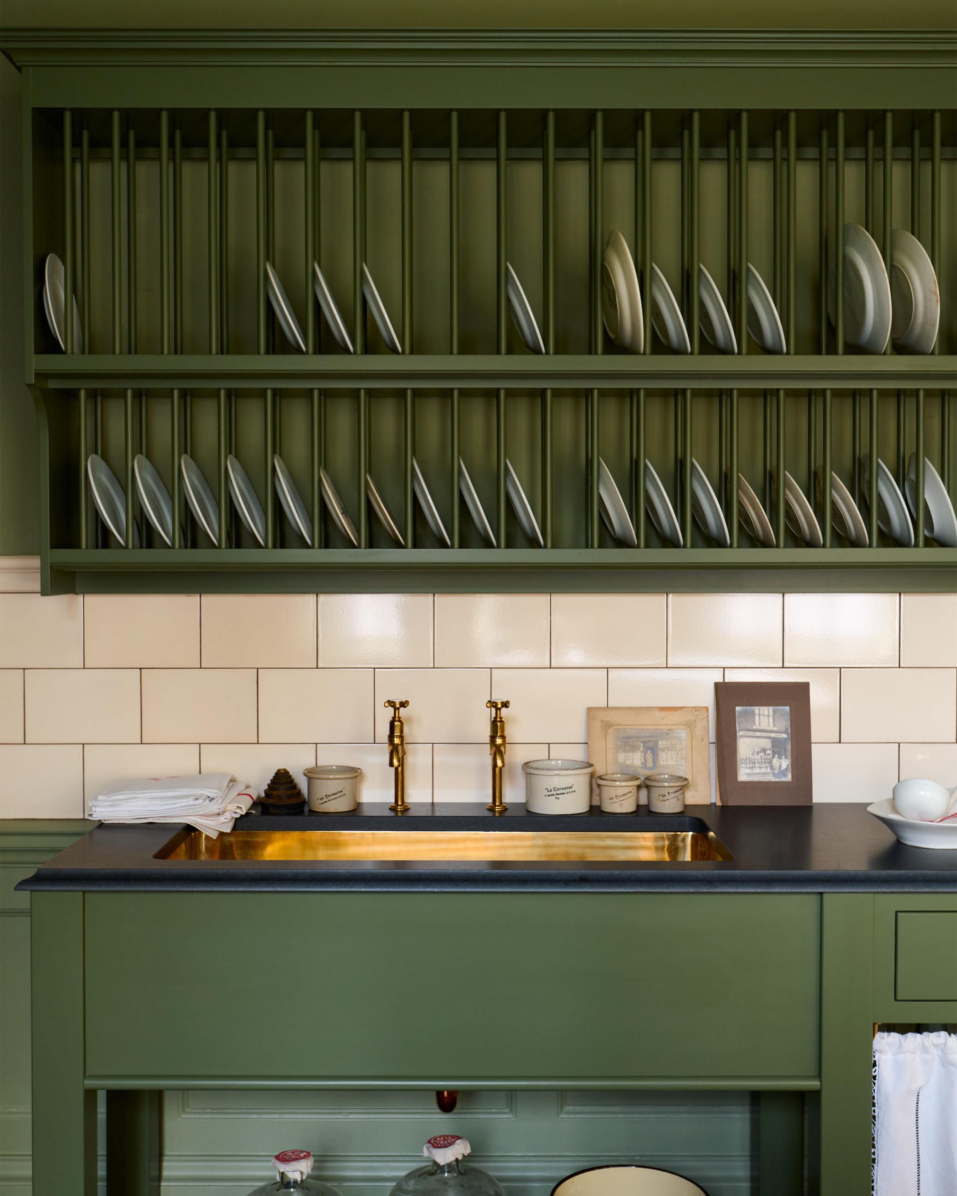 A close-up straight-on view of a mossy green painted Victorian-style sink cupboard with a polished brass sink, aged brass bibcock taps and cream subway wall tiles below a traditional wall-mounted plate rack.
