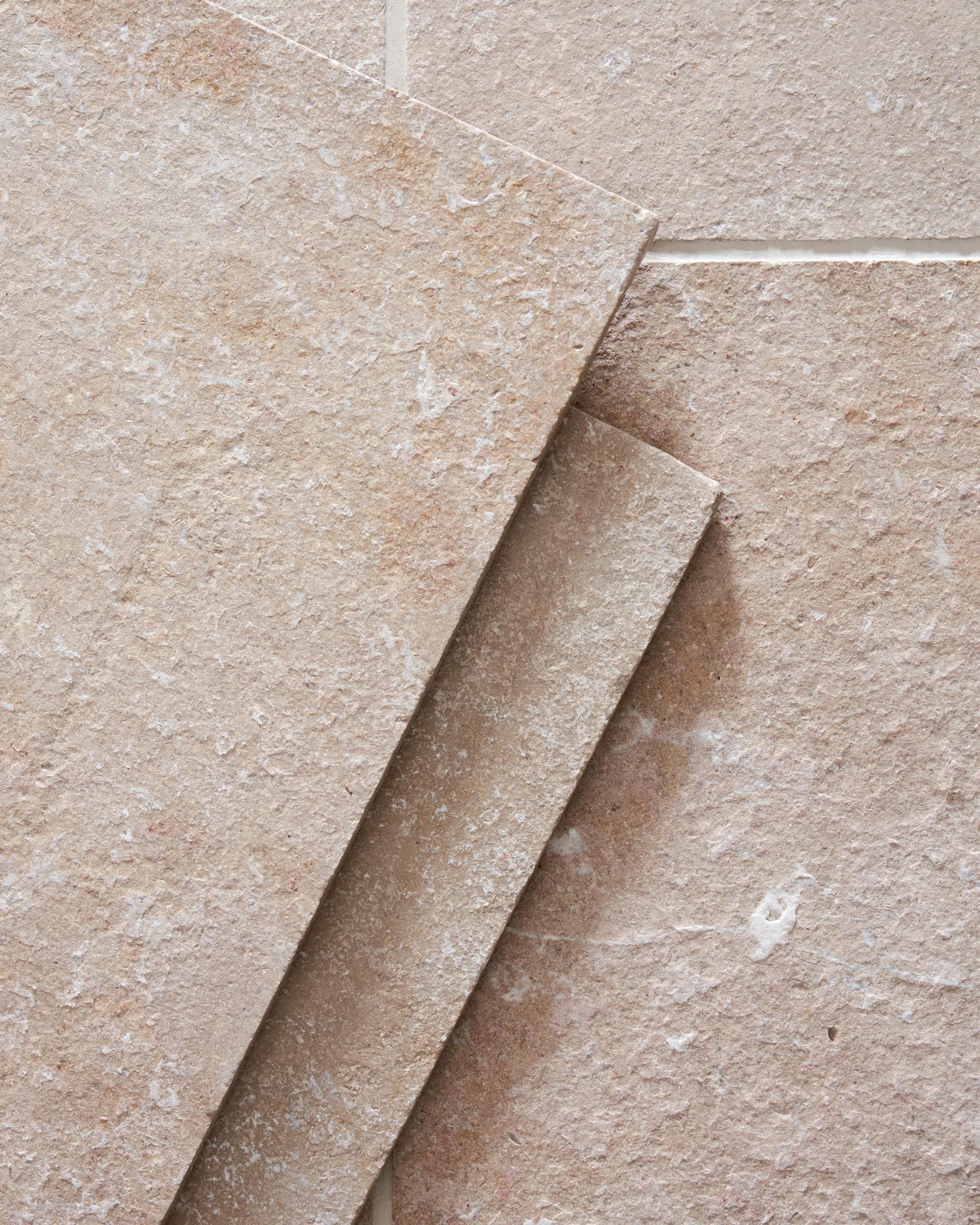 A close-up overhead view of a soft creamy pink coloured limestone floor with a lightly textured surface, with two loose tiles stacked in one corner.