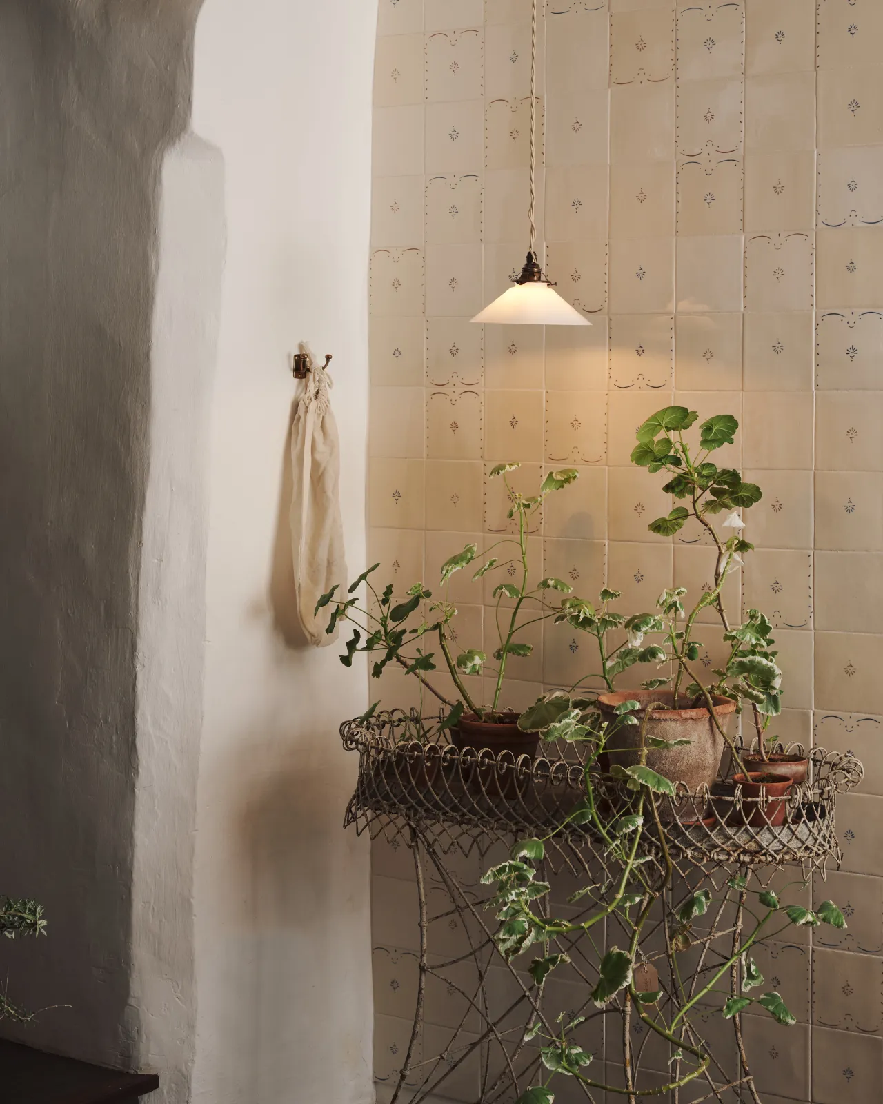 A small pendant light with translucent milk lamp shade in front of a wall filled with ditsy delft tiles pattern, with a console table with potted plants.